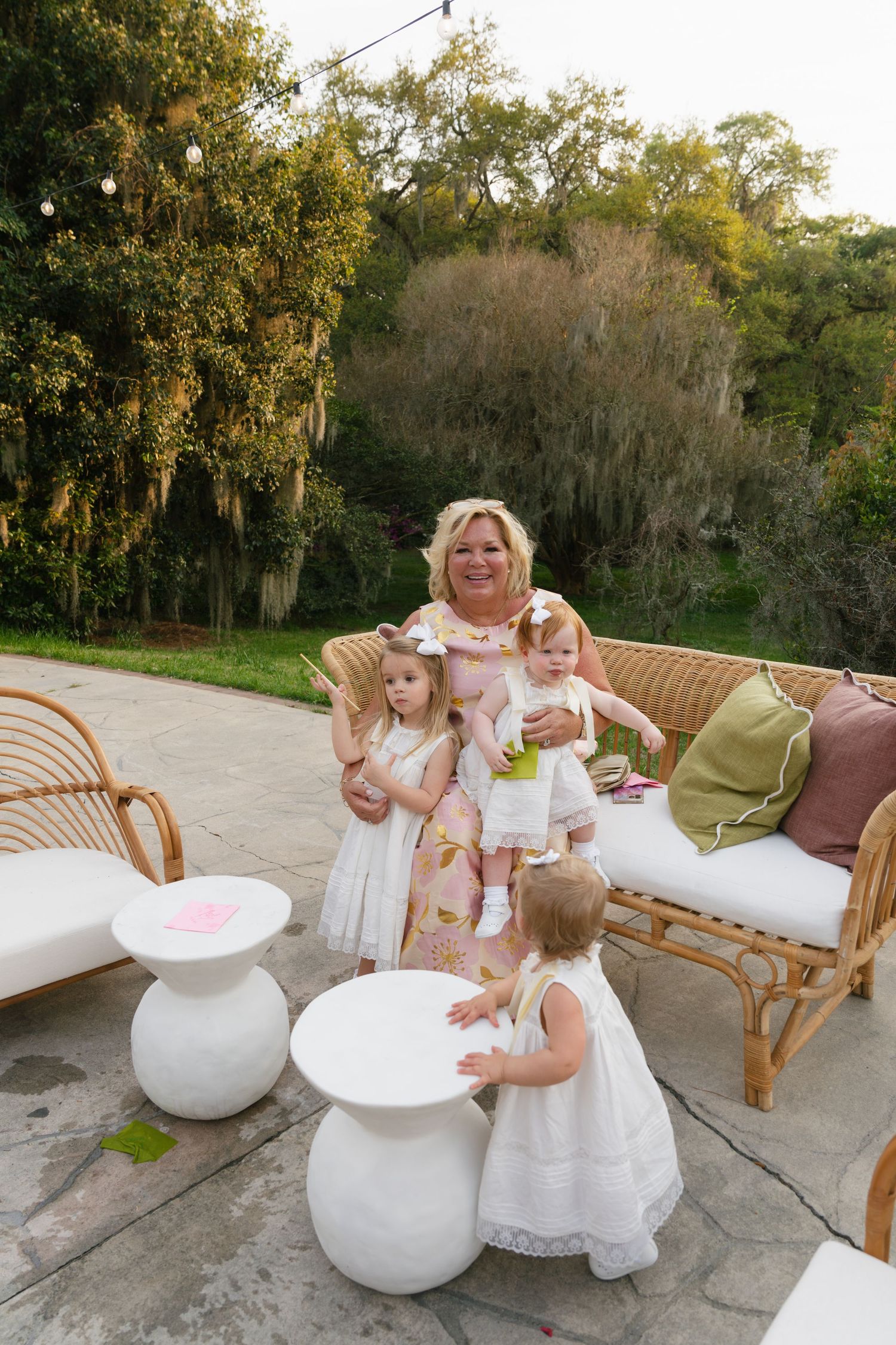 Grandmother poses with three young girls in white dresses on a patio with rattan furniture and lush garden backdrop.
