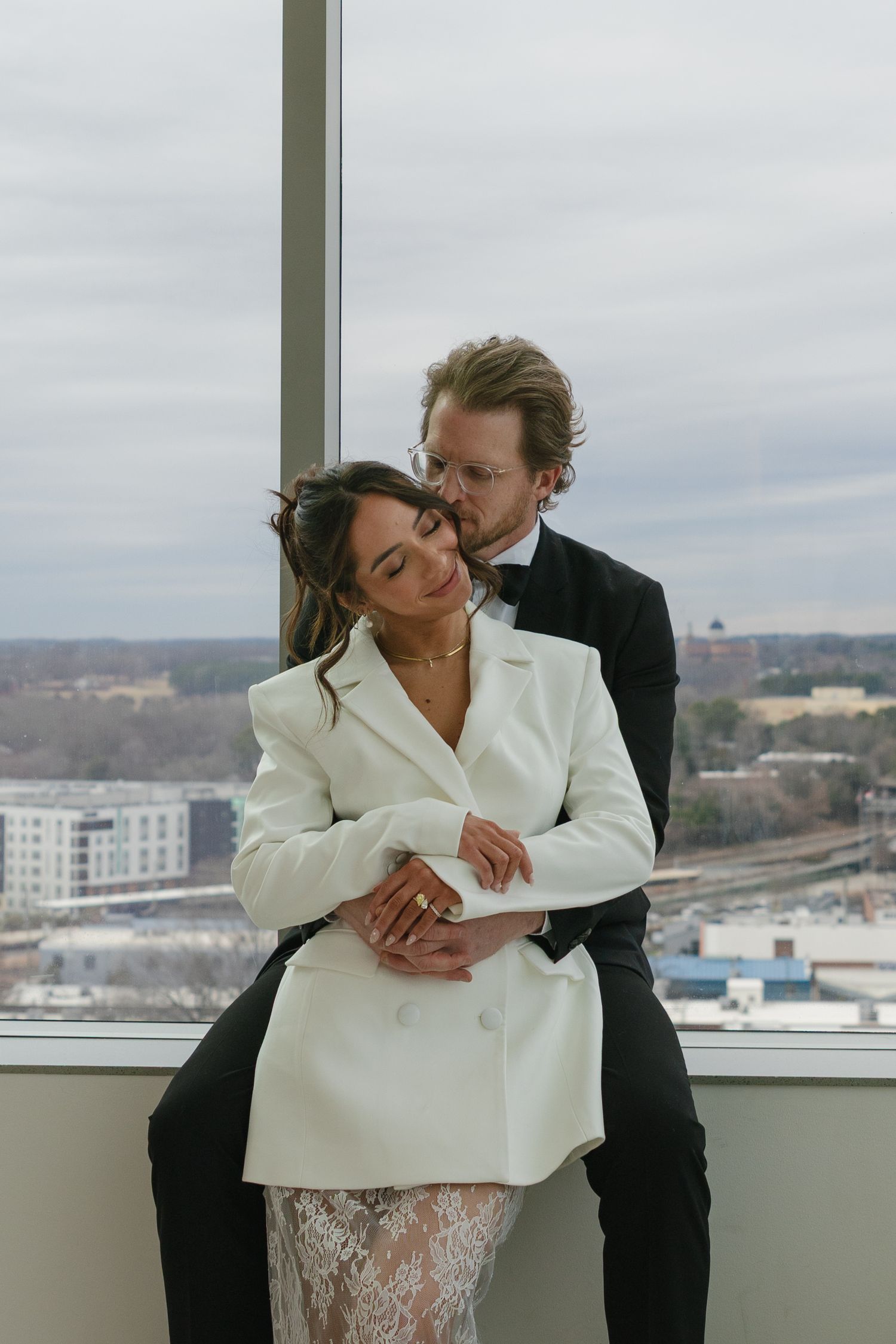A couple in wedding attire embraces by a high-rise window, bride in white blazer dress, groom in black tuxedo kissing her cheek.