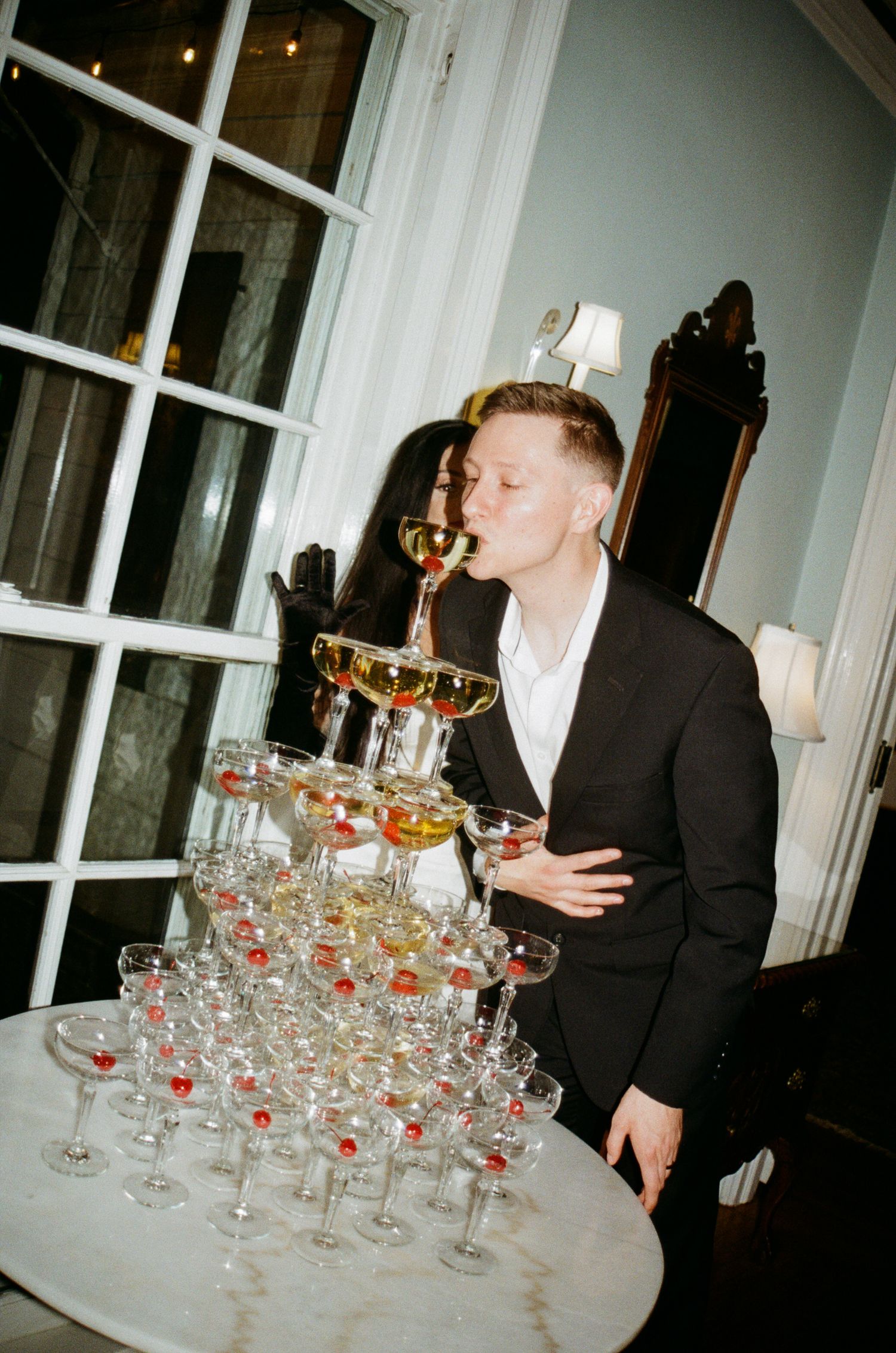 Man in black suit drinking champagne from a coupe glass beside a pyramid tower of glasses at an elegant party.