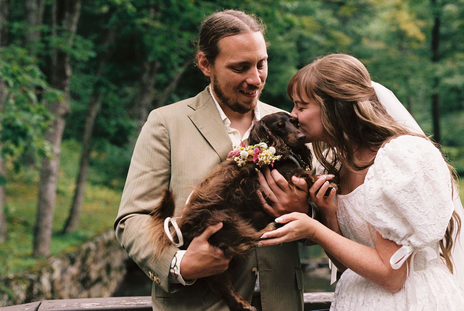 A couple in wedding attire shares a joyful moment while holding a brown cat in a wooded outdoor setting.