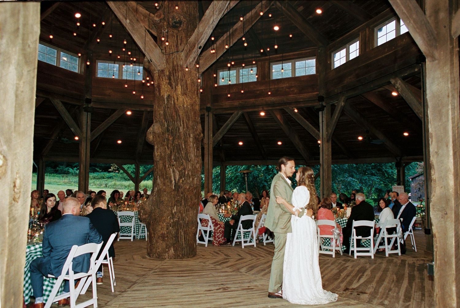 A couple shares their first dance in a rustic wooden barn venue surrounded by guests seated on white folding chairs.