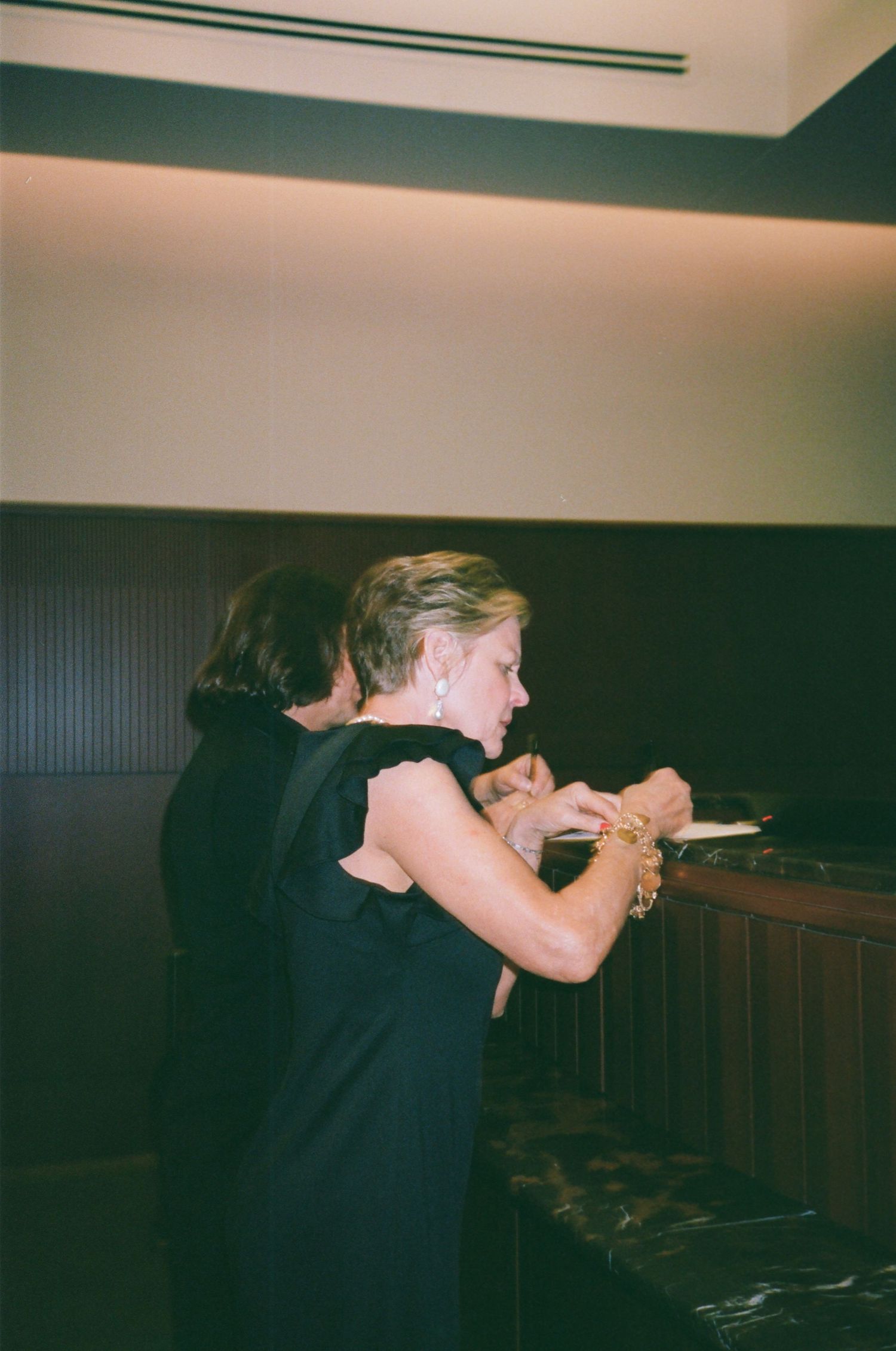Two women in black dresses at a formal event, one wearing gold bracelets, standing near a dark marble counter.