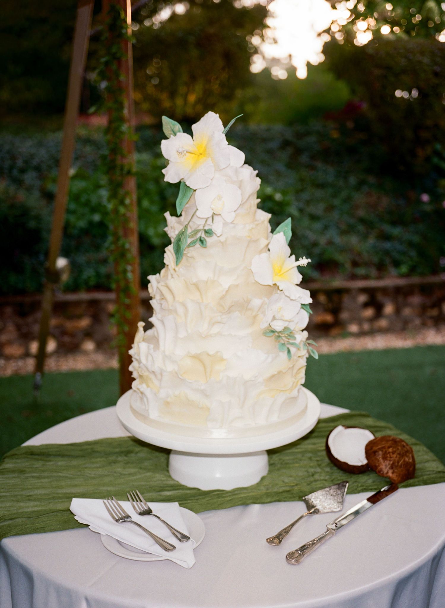 An elegant white wedding cake with delicate floral decorations sits on a table in an outdoor garden setting.