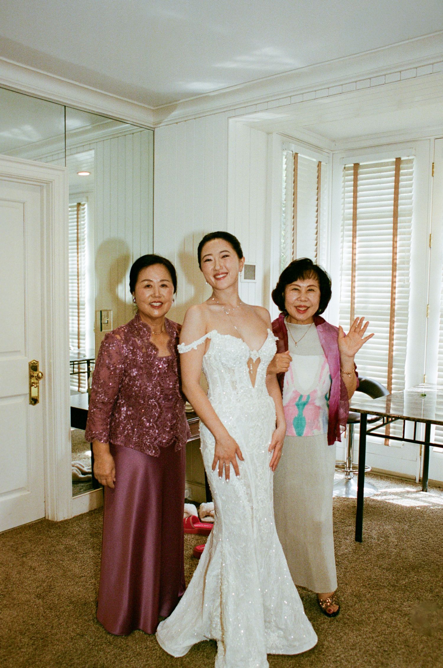 A group poses joyfully in a bright hotel room with white walls, brown carpeting and window blinds.