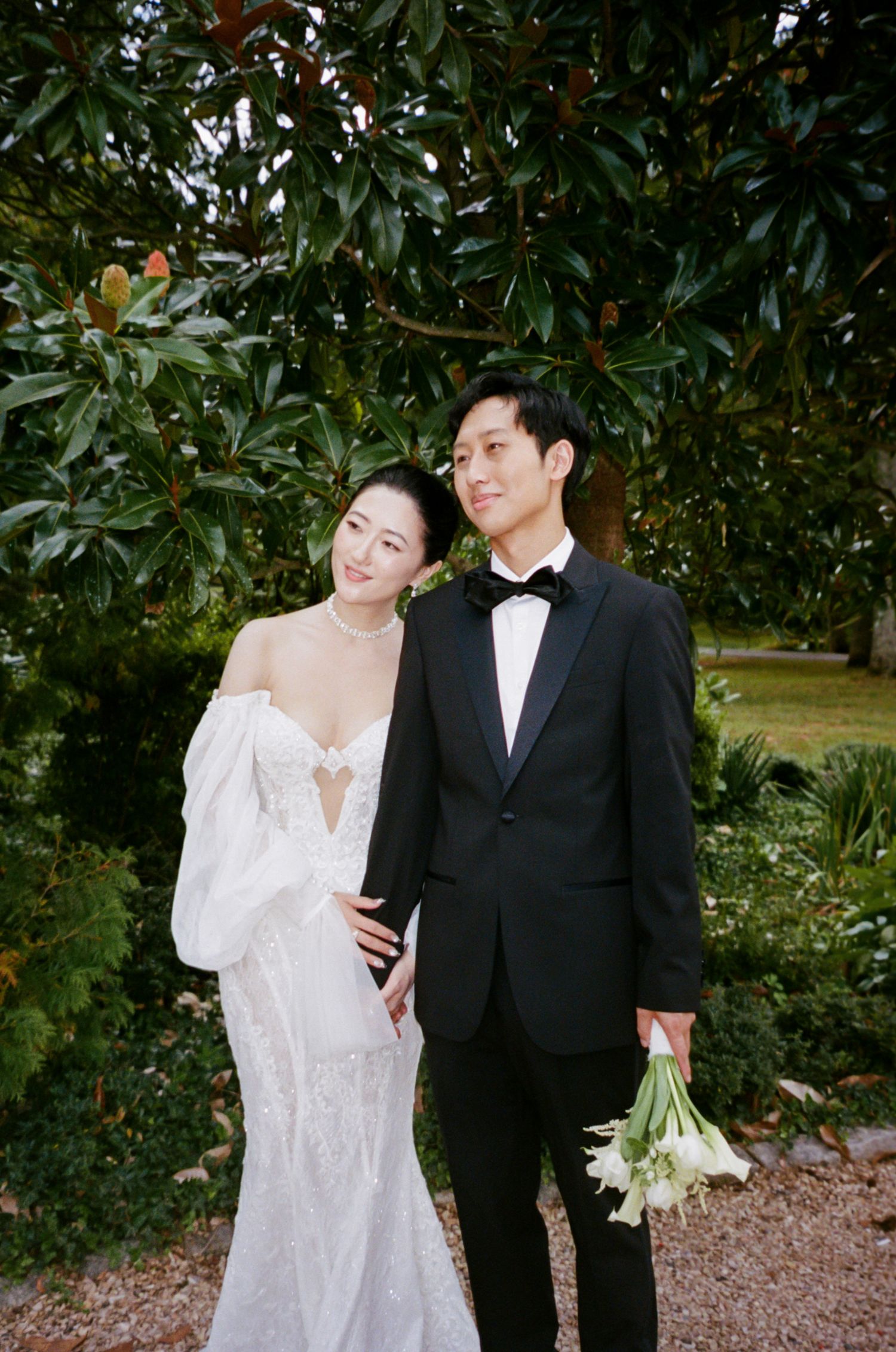 A couple poses together outdoors in formal wedding attire surrounded by lush greenery and trees.