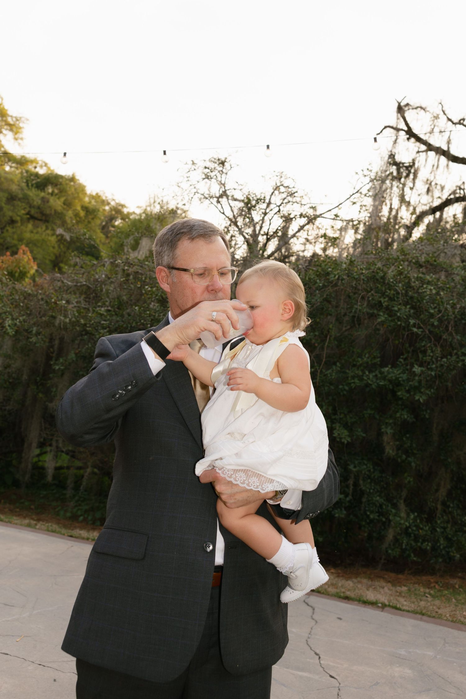 Man in dark suit holding a baby girl in white dress, feeding her from a bottle outdoors near trees.