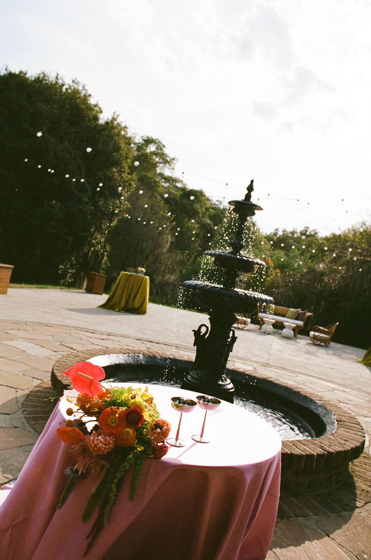 Outdoor wedding reception with colorful floral arrangement and wine glasses on a pink-draped table beside an ornate black fountain.