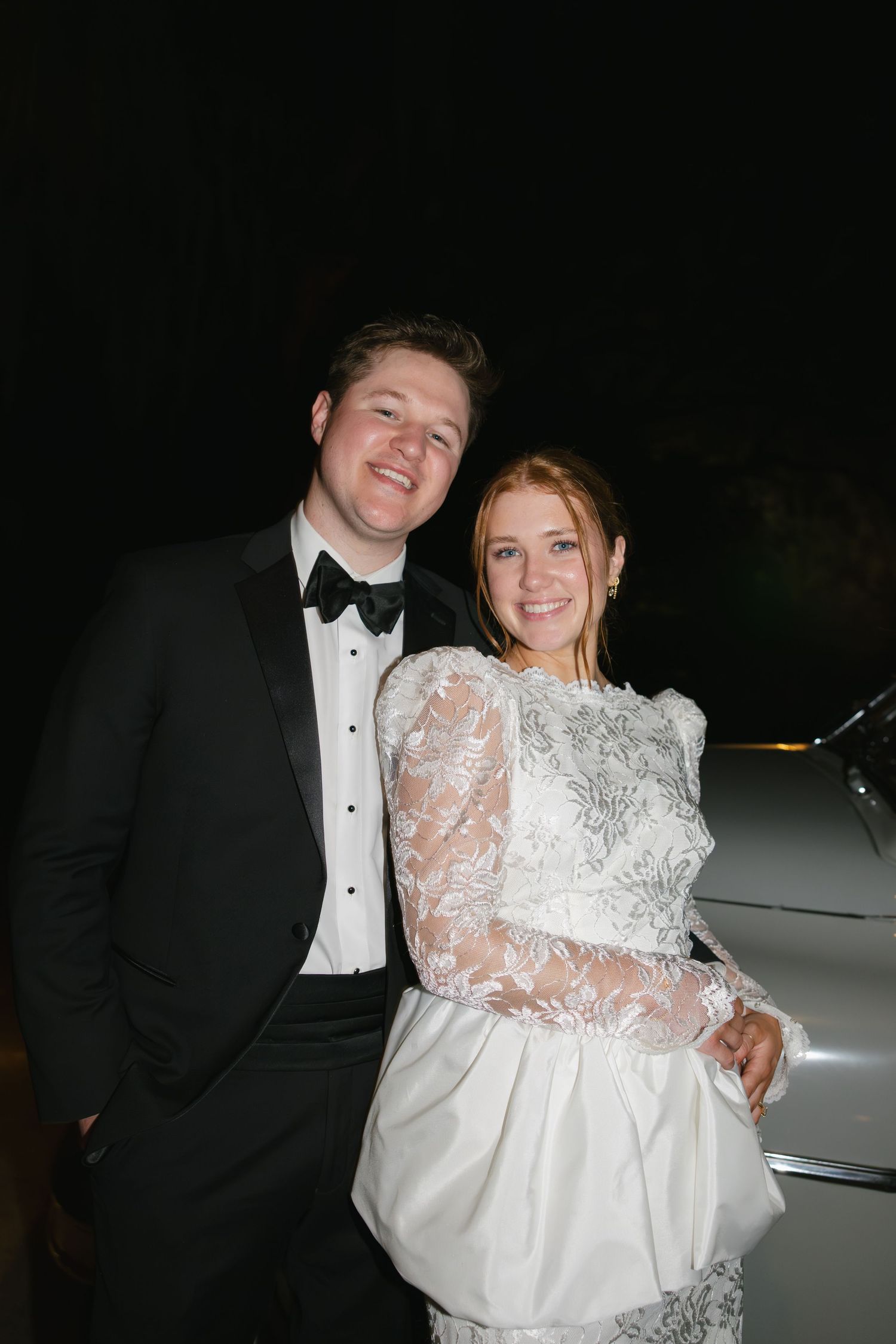 A smiling couple in wedding attire, groom in tuxedo and bride in lace dress, posing together at night.