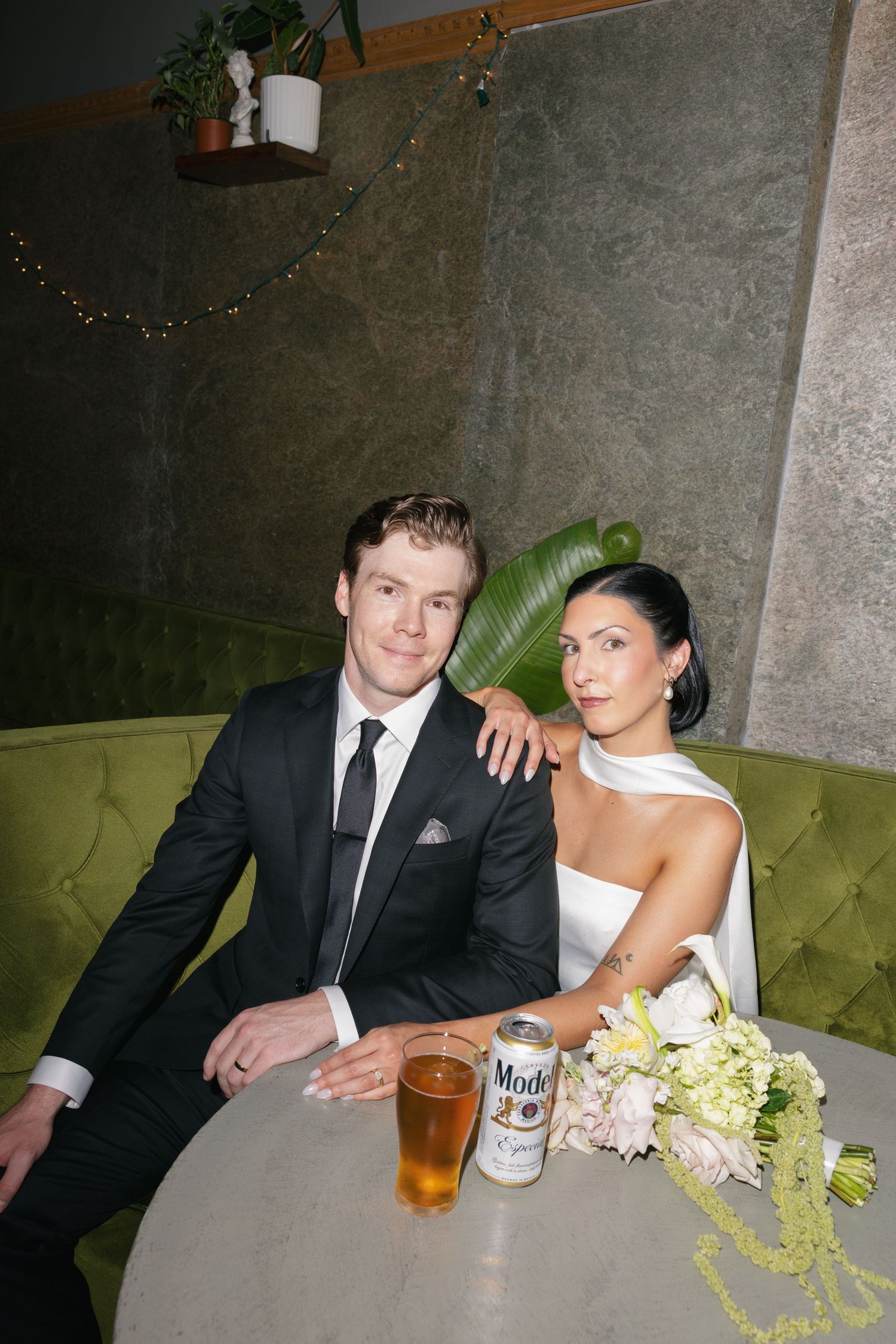Bride in a white dress and groom in a black suit sit together on a green velvet booth with a bouquet, a Modelo can and a beer on the table.