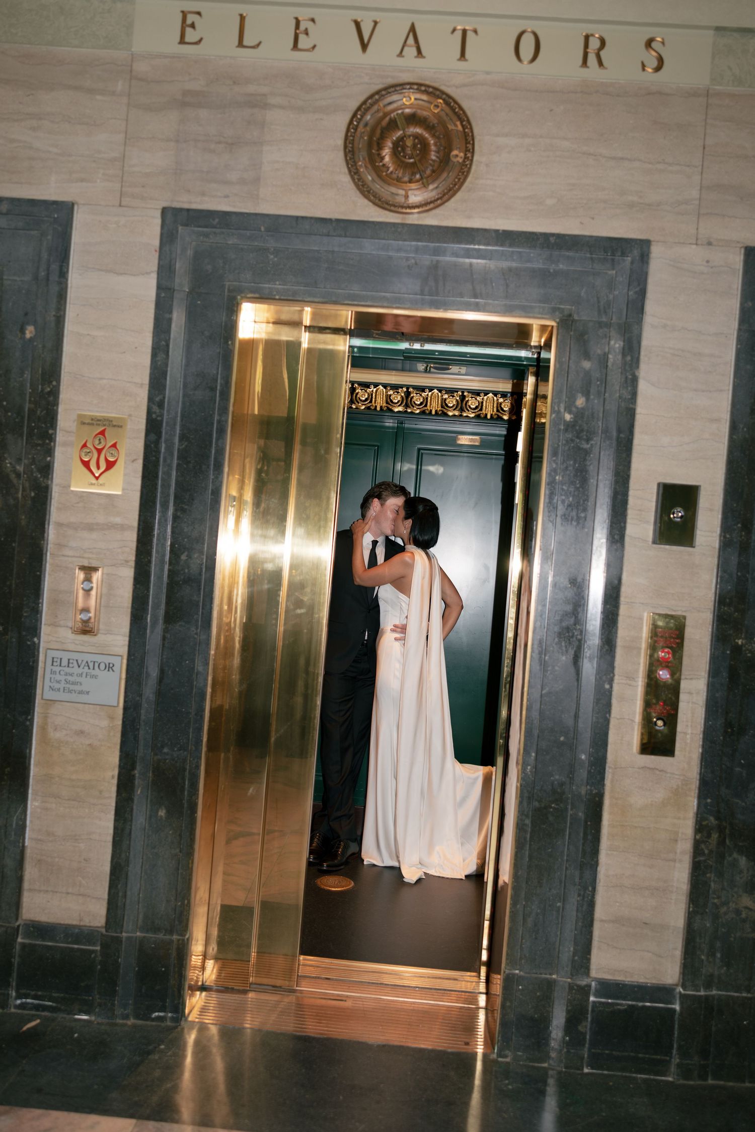 Bride in a white gown and groom in a dark suit kiss inside a gold elevator beneath an ornate marble Elevators sign.