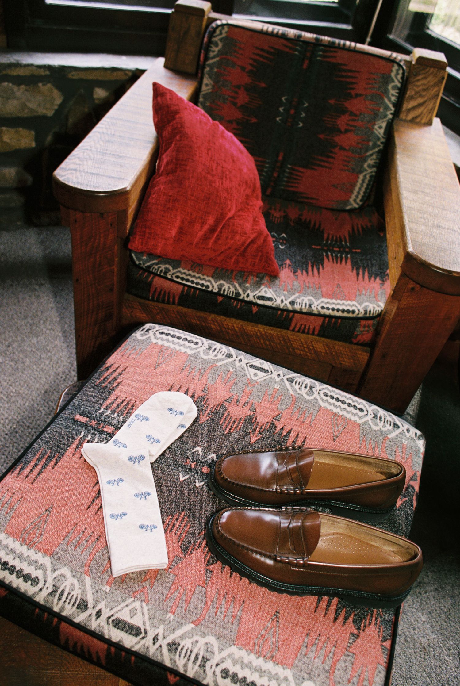 Brown leather loafers rest on a red and black patterned rug in a dimly lit room.