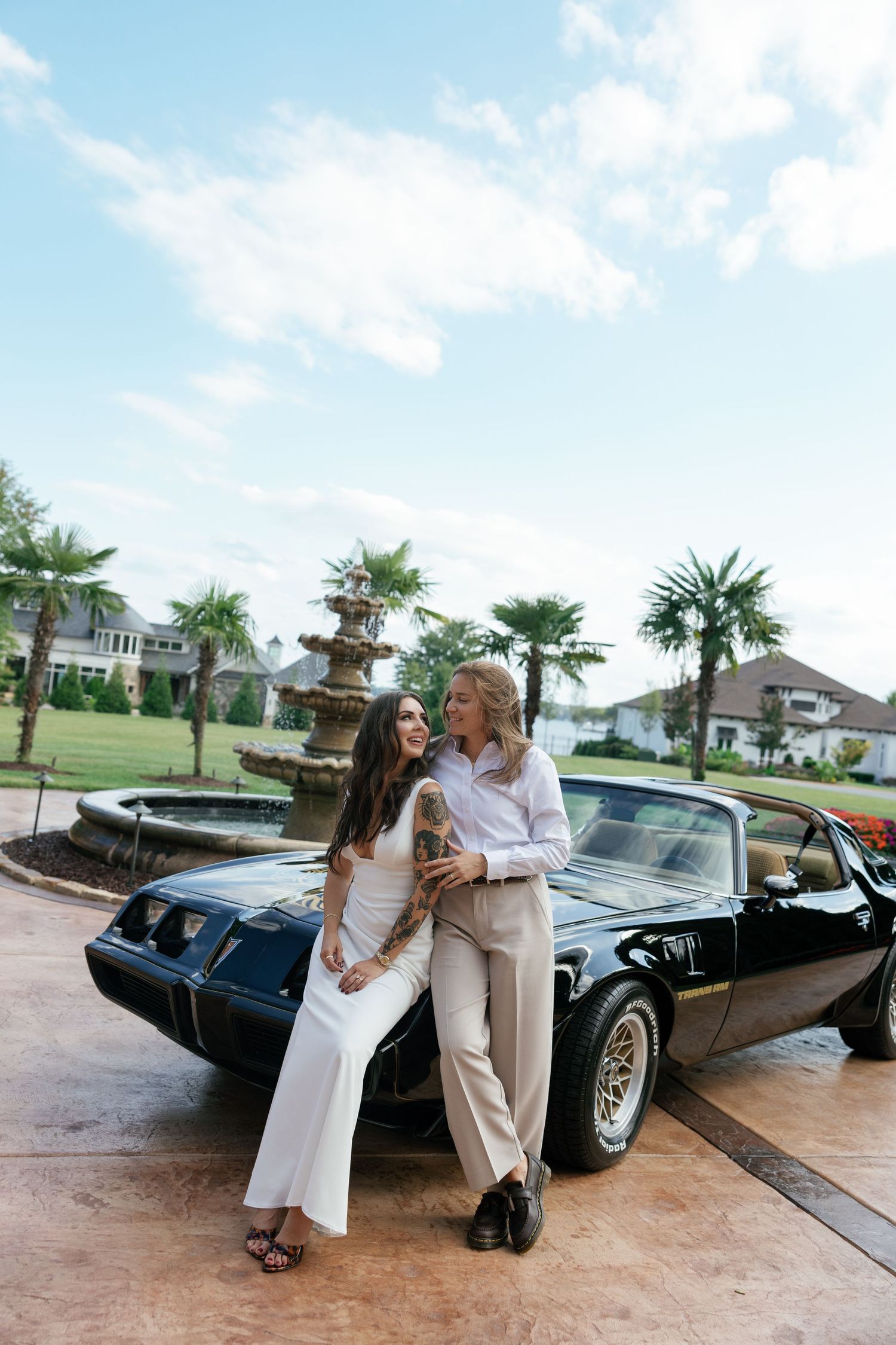A classic black vintage car serves as backdrop for a stylish couple posing in front of palm trees.
