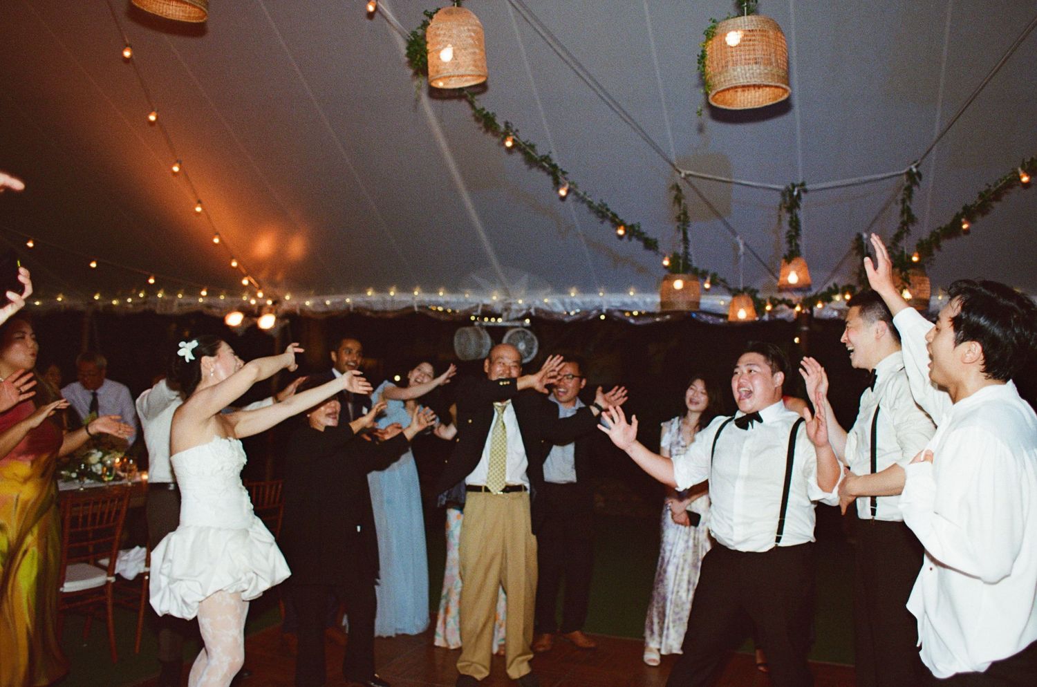 Wedding reception guests dancing under a tent decorated with string lights and hanging lanterns.