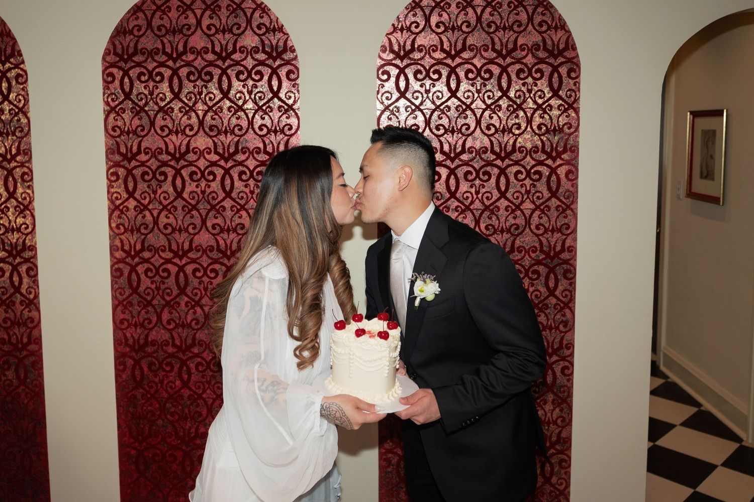 A wedding couple stands against a deep red damask wallpaper background in an elegant indoor setting.