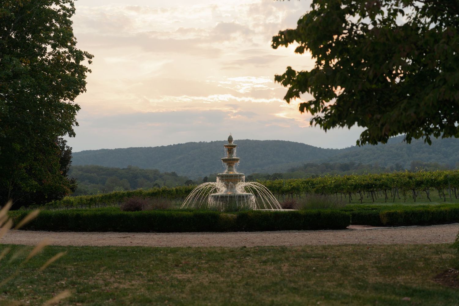 Scenic panoramic view of rolling hills and vineyards at sunset with a decorative stone fountain in the foreground.