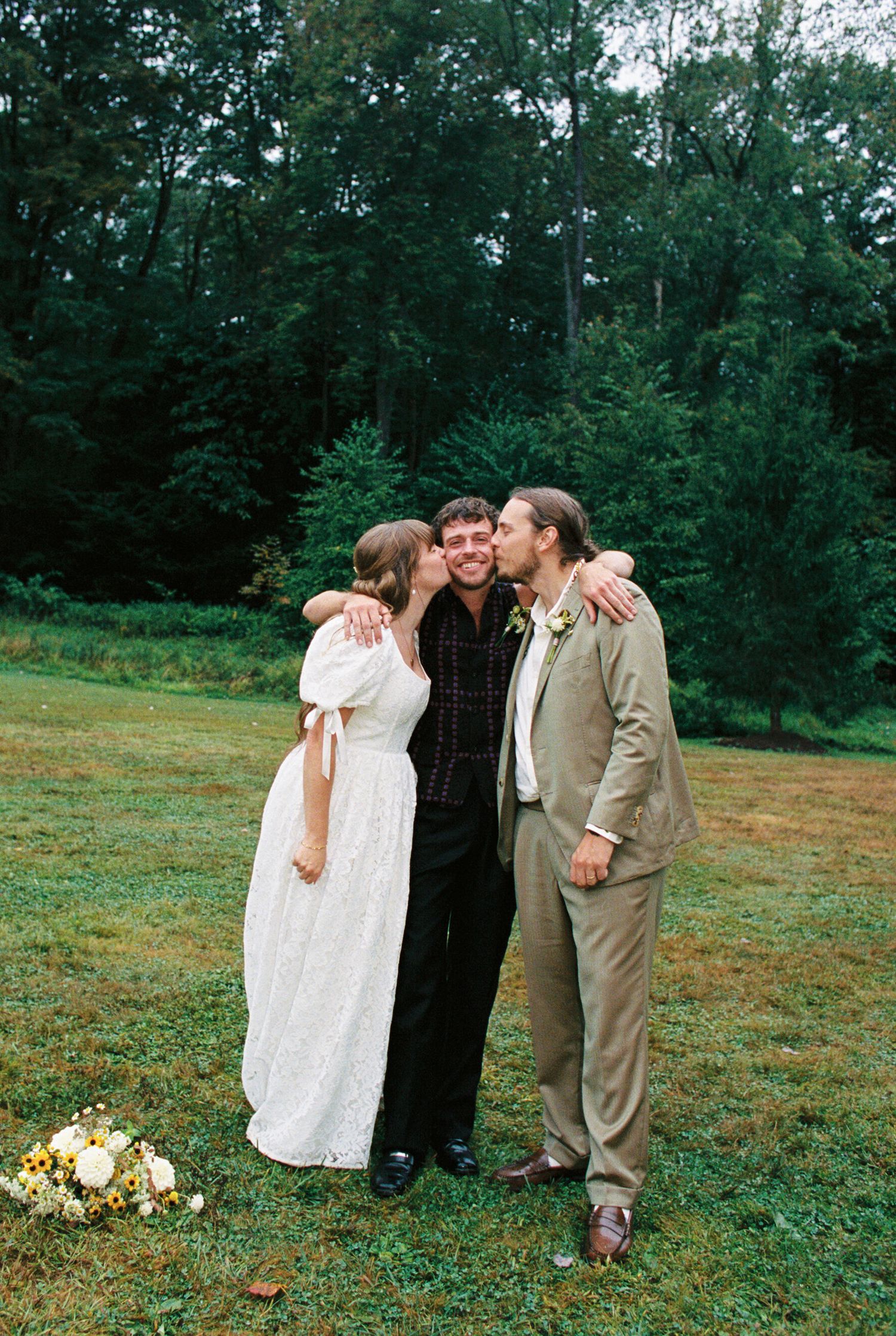 A romantic outdoor wedding moment captured on a grassy lawn with trees in the background and a small floral bouquet nearby.