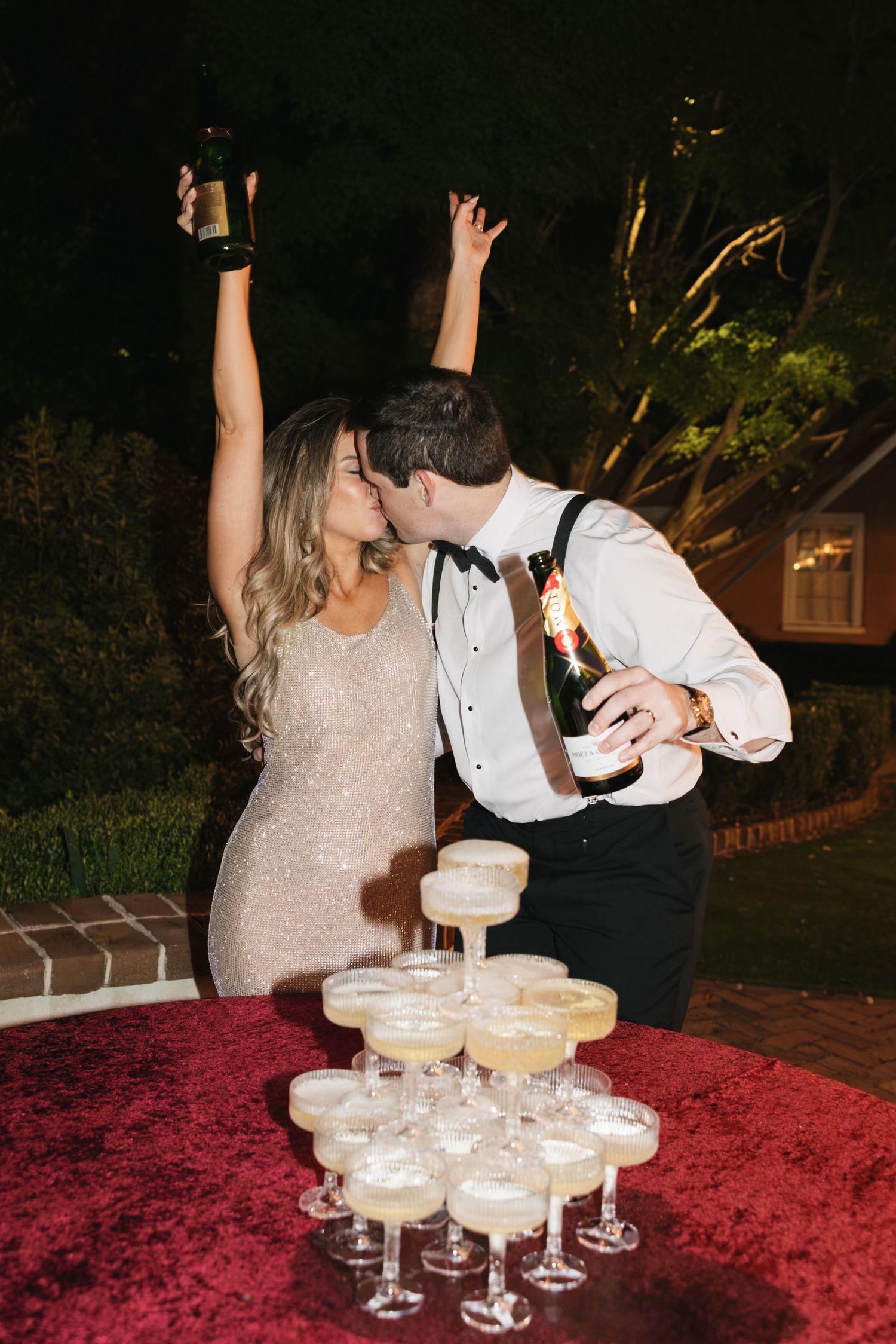 Couple celebrates joyfully with champagne tower at elegant party, woman in sparkly dress raising glass beside stacked coupe glasses.