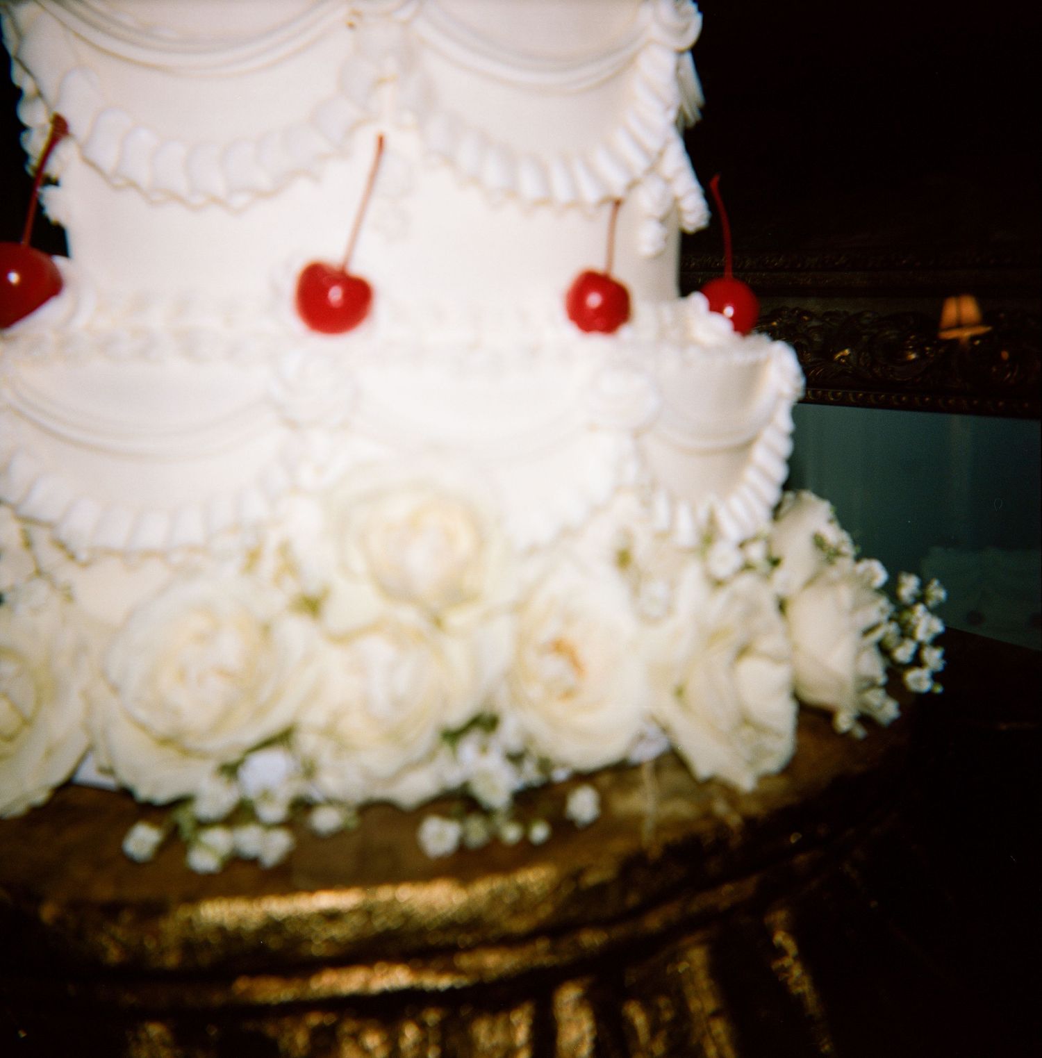 White tiered wedding cake with piped scalloped frosting, red cherries on top, and fresh cream roses and baby's breath on a gold stand.