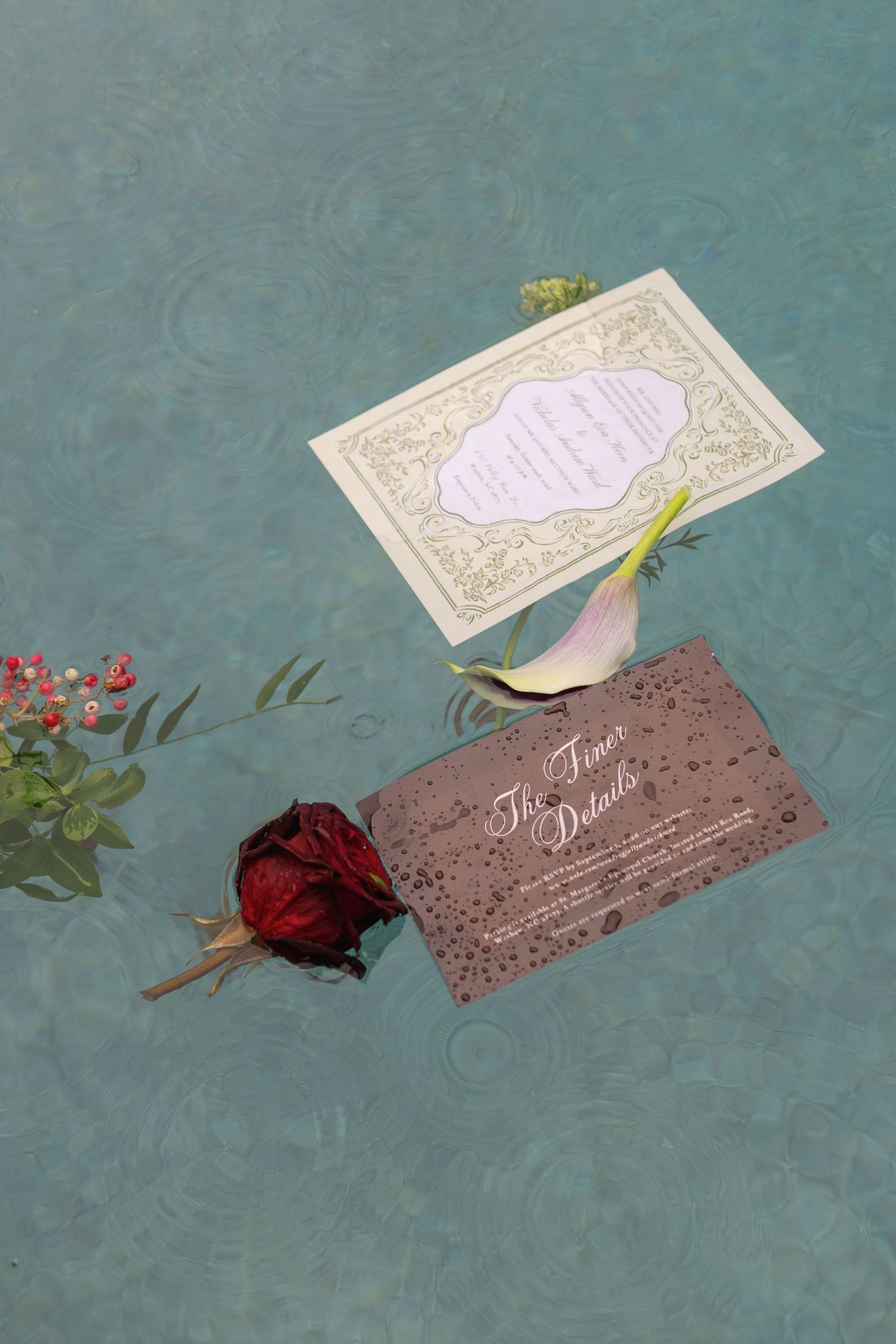 Ornate cream wedding invitation and a brown details card float on turquoise water beside a red rose, calla lily and berry sprig.