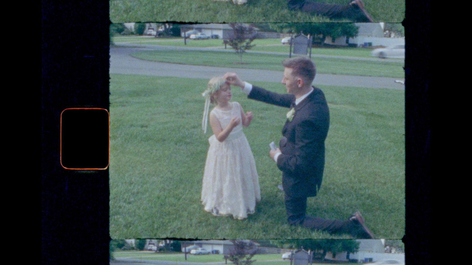 Vintage film still of a man in a dark suit kneeling on a green lawn, adjusting a young girl's floral headpiece.
