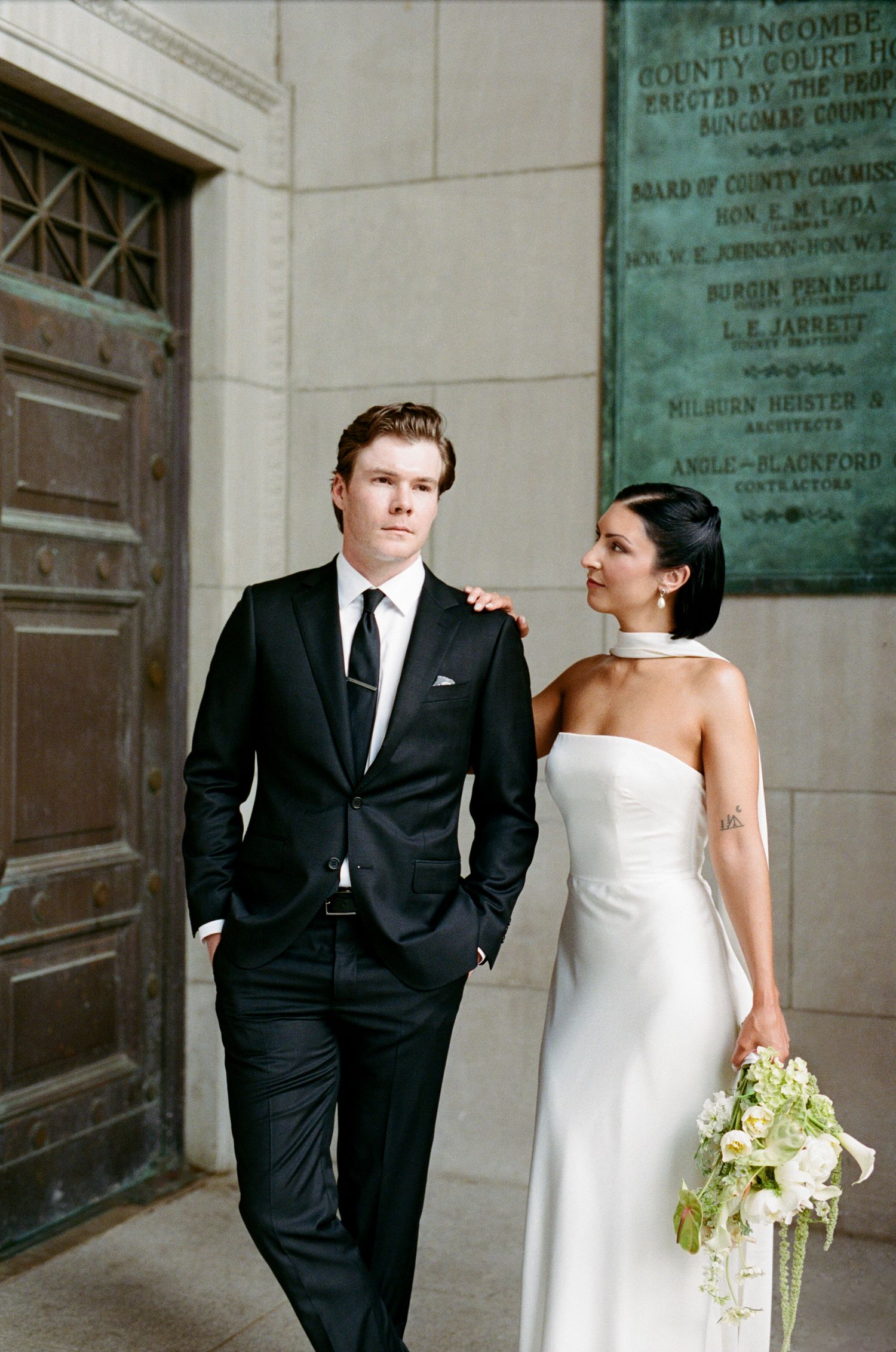 Bride in a strapless white gown holding a bouquet stands beside a groom in a black suit before a bronze door and plaque.