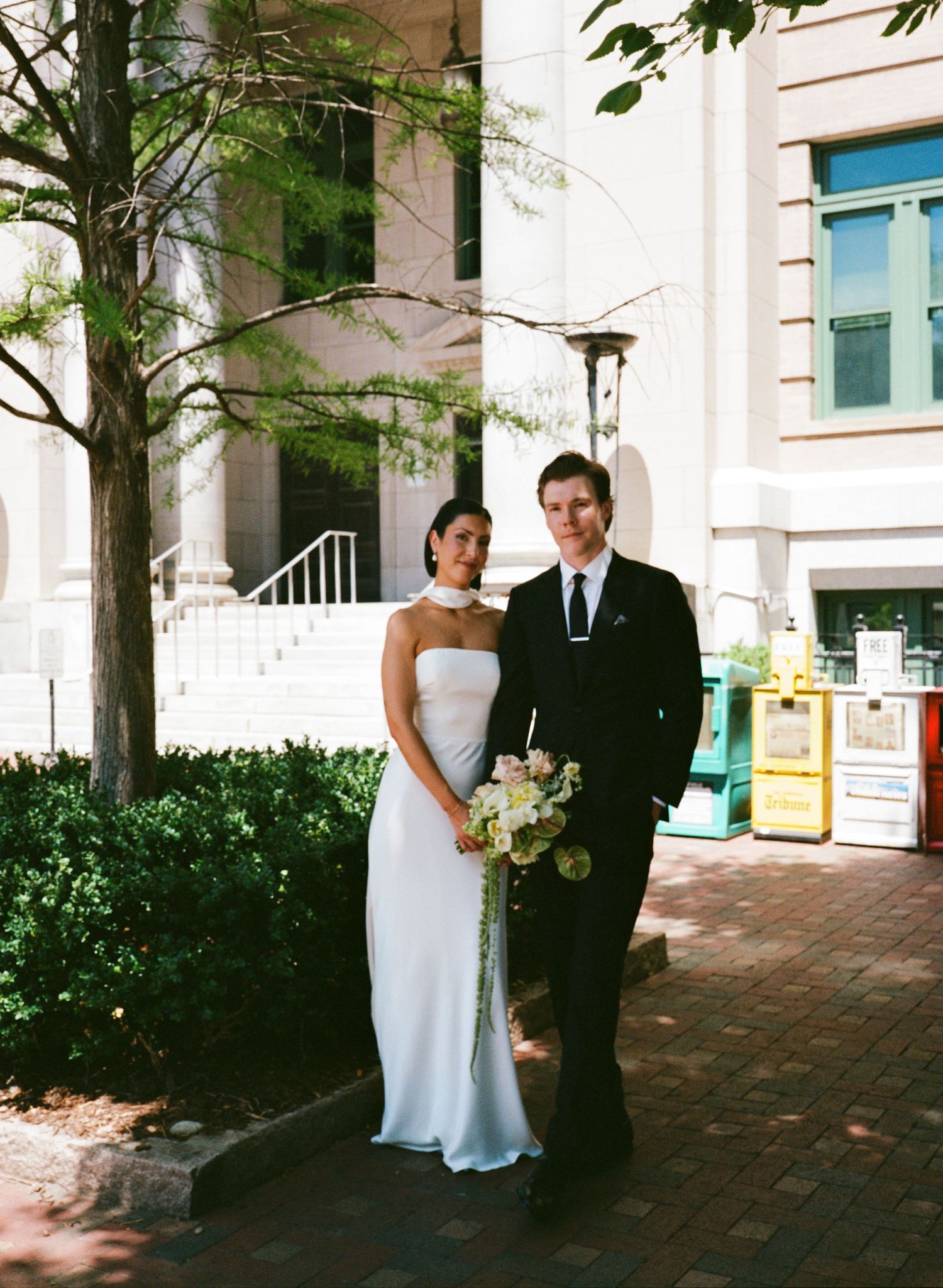 Bride in a white strapless gown holding a pastel bouquet stands beside a groom in a black suit outside a stone building.