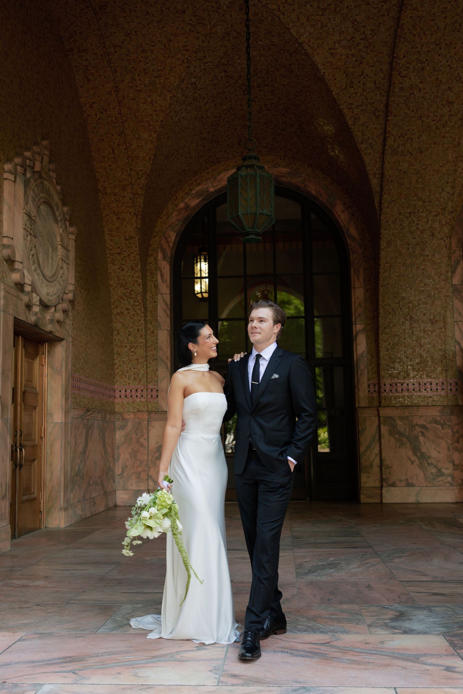 Bride in a strapless white gown holding a bouquet smiles up at the groom in a dark suit beneath an arched stone doorway.