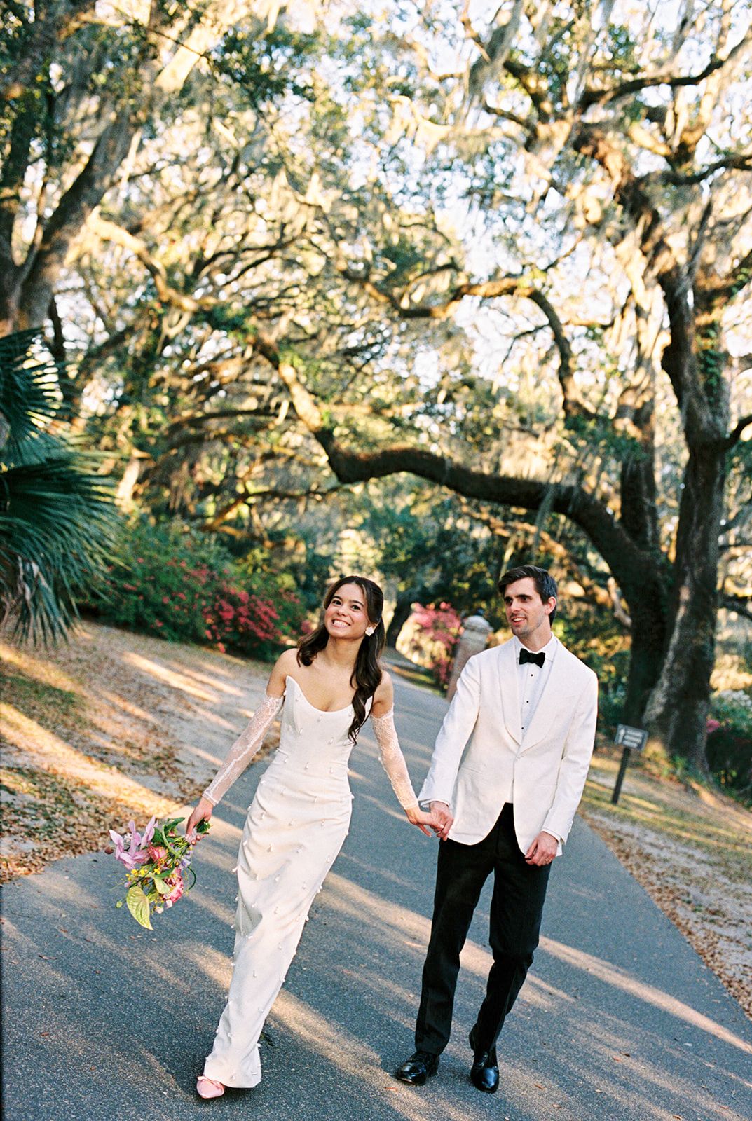 A couple in wedding attire strolls along a tree-lined path during a golden sunset photo session.