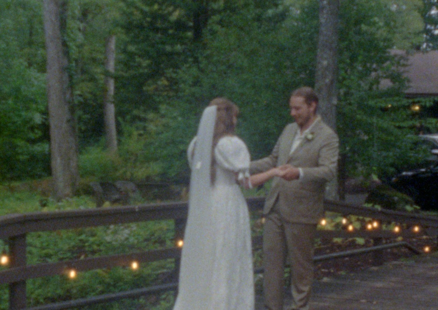 A wedding ceremony takes place on a wooded path decorated with glowing string lights at dusk.