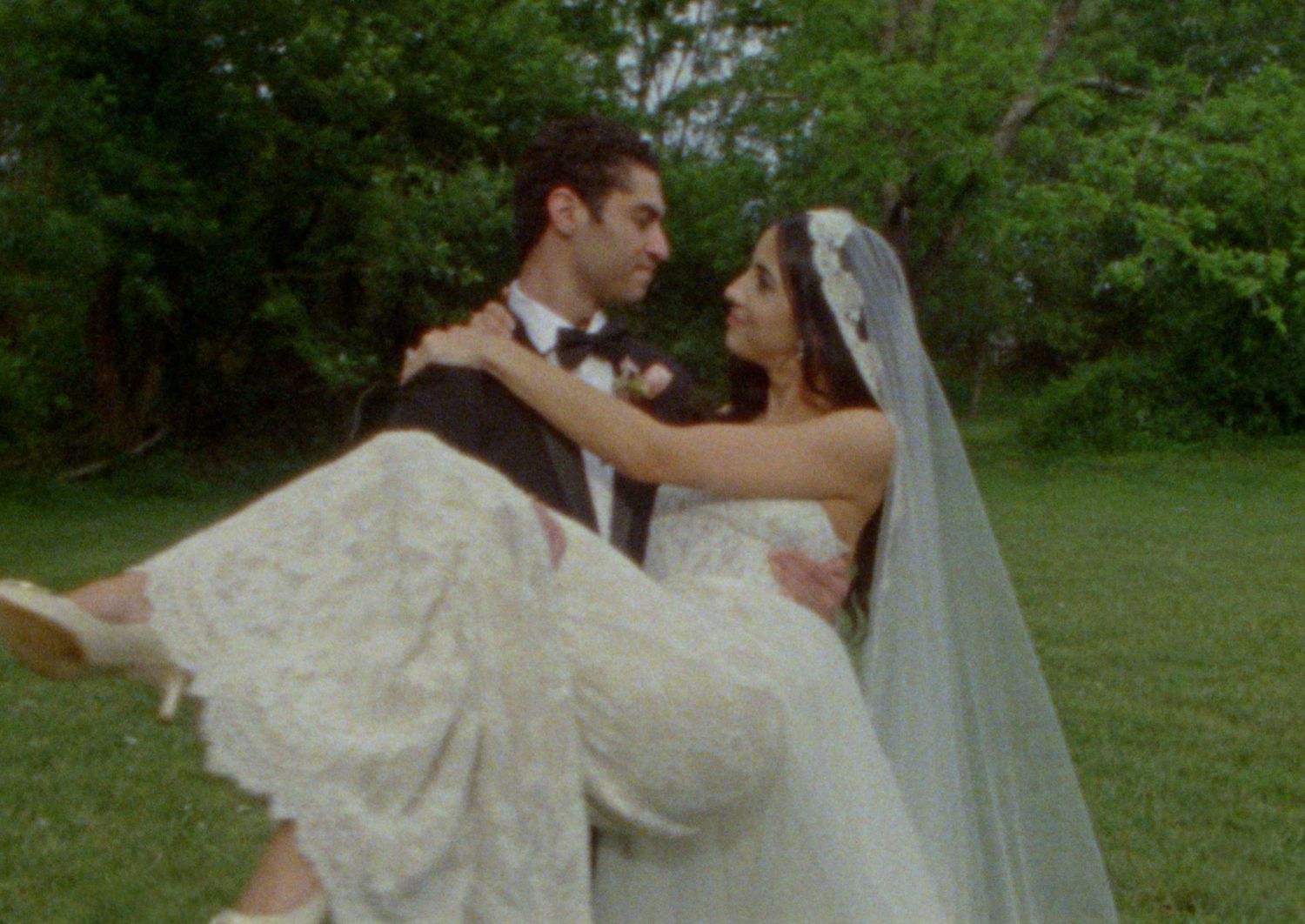 Groom carrying bride in white lace wedding dress and veil outdoors on green lawn.
