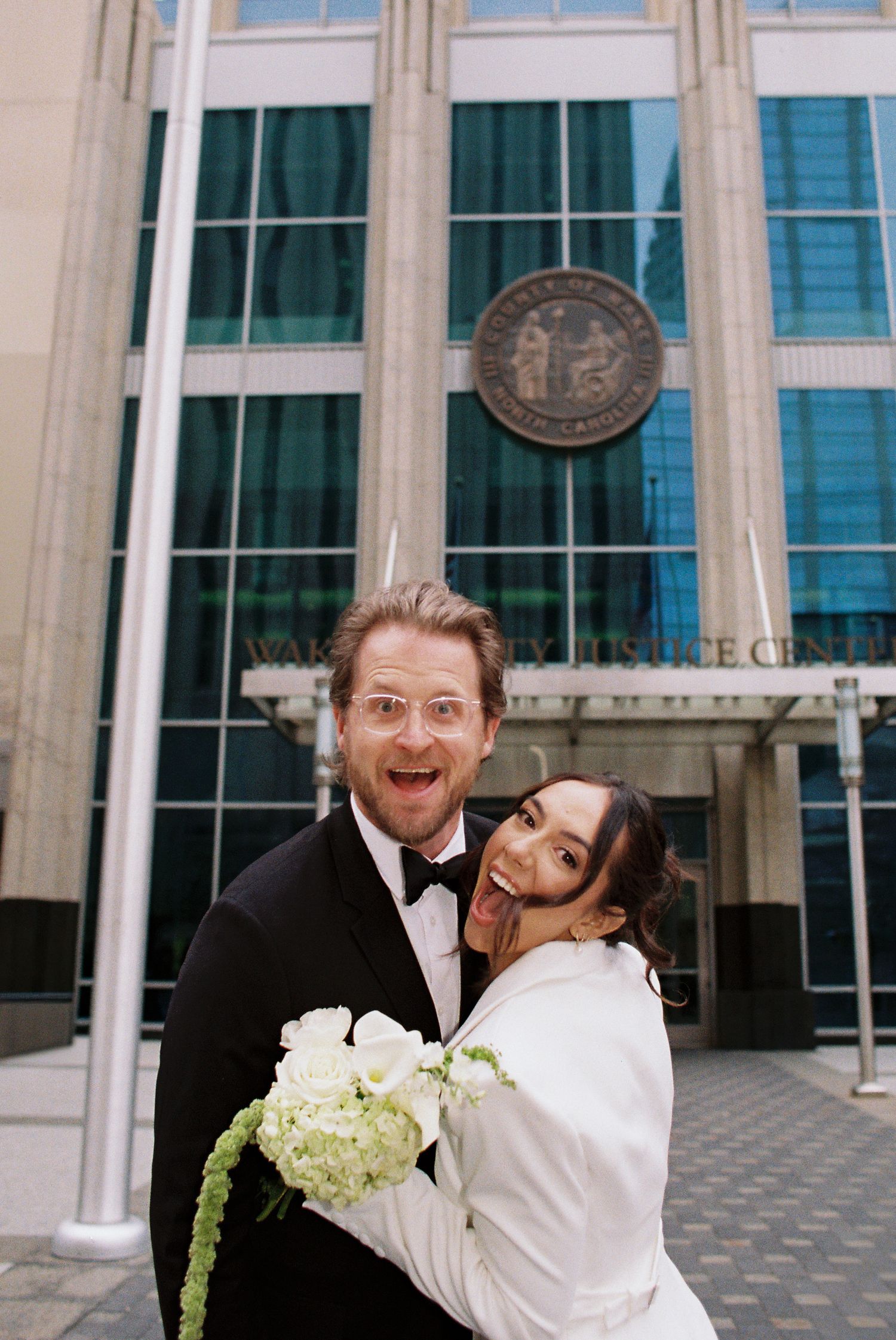 Joyful newlyweds posing outside a courthouse, bride in white holding bouquet, groom in tuxedo, celebrating civil wedding ceremony.
