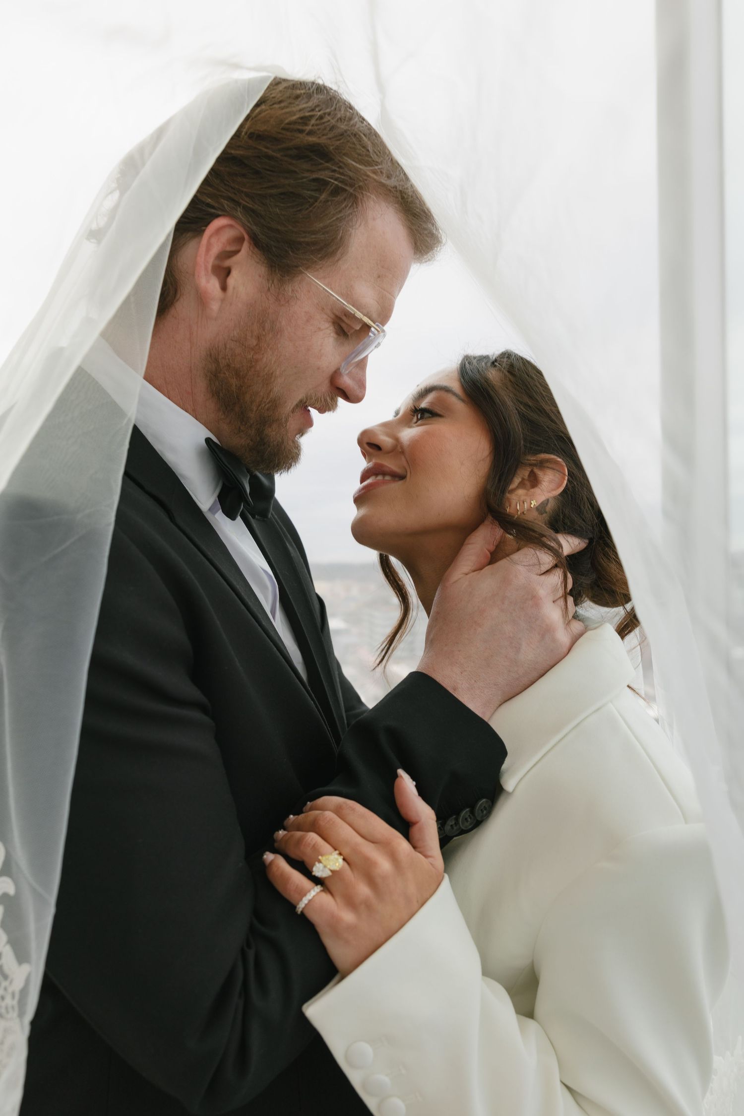 Romantic wedding couple sharing an intimate moment under a veil, groom in black tuxedo, bride in white dress with gold rings.