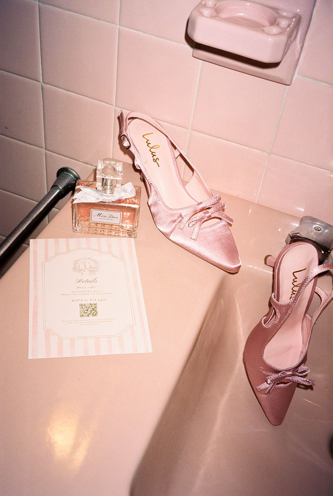 Pink satin heels with bow detail beside Miss Dior perfume and invitation card on a pink bathroom ledge.
