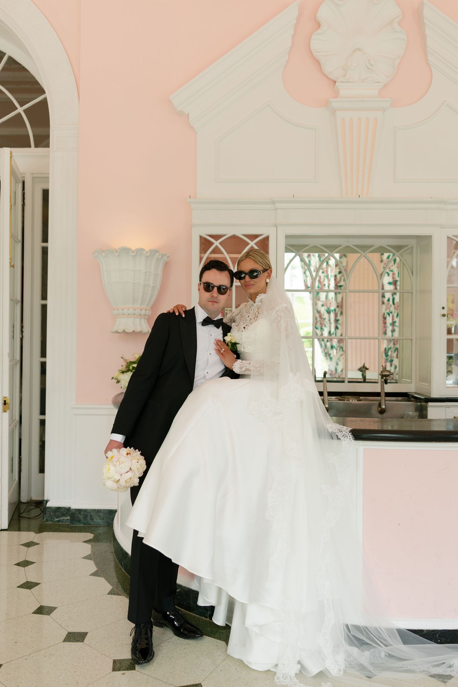 Bride and groom in sunglasses pose elegantly in a pink venue, she in a white gown with veil, he in a black tuxedo.