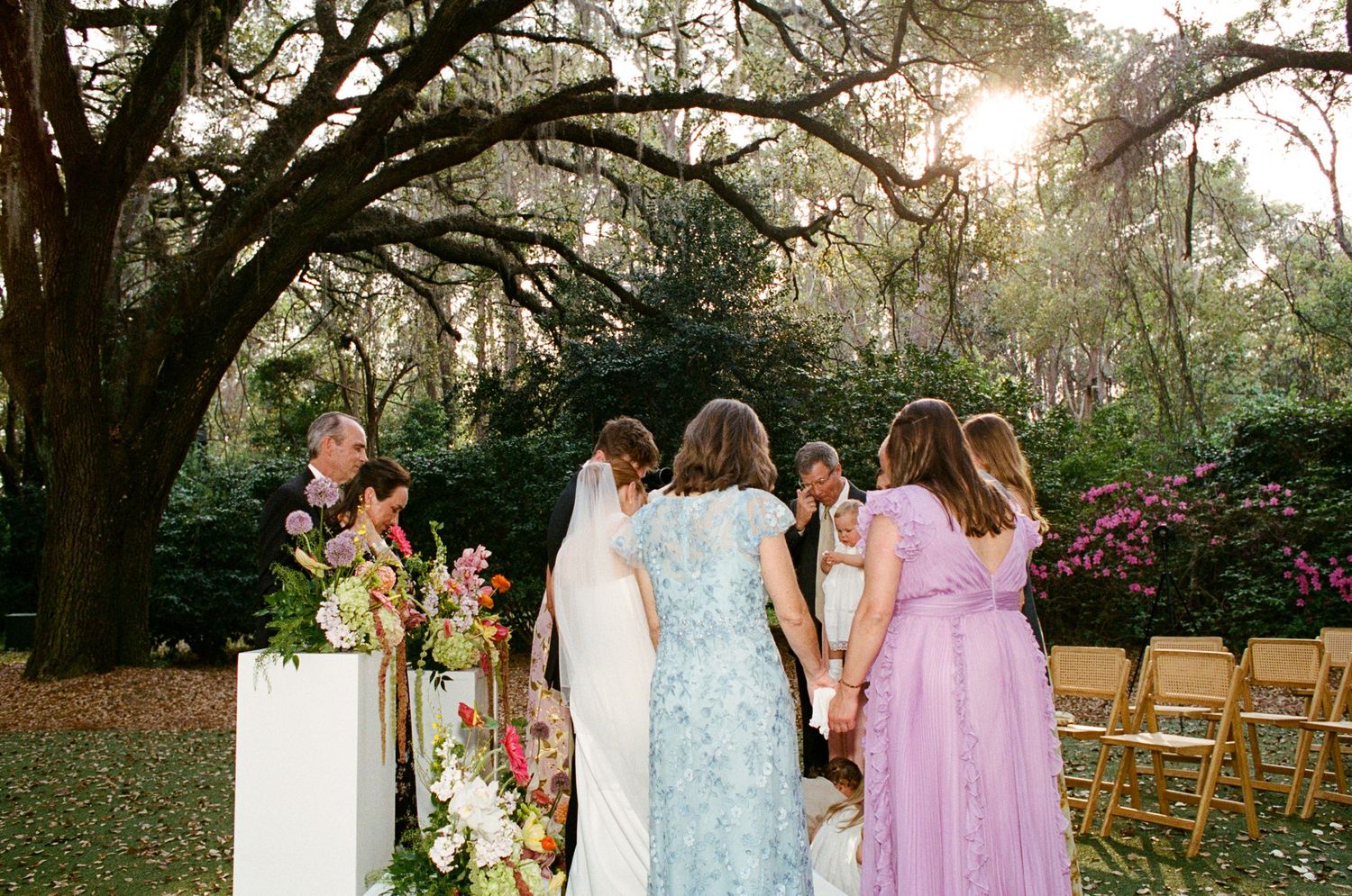 Bride in white veil with bridesmaids in colorful dresses at outdoor wedding ceremony under oak trees.