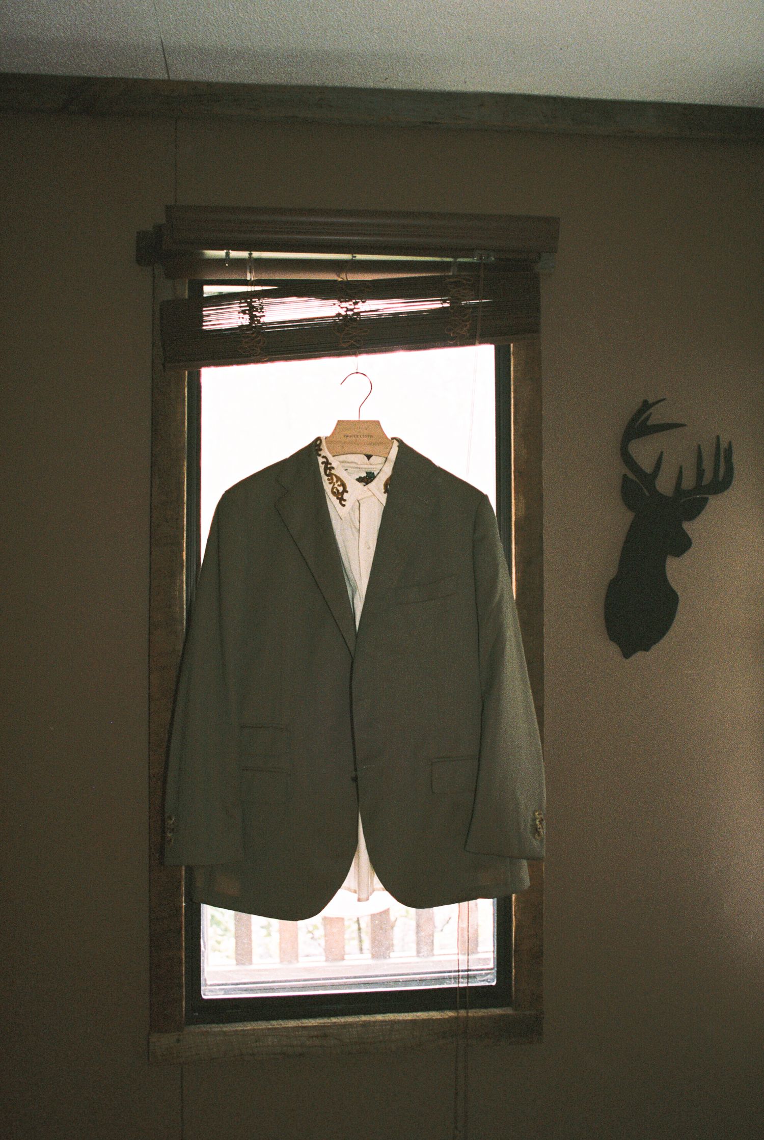 A green blazer jacket hangs in front of a window with a deer head silhouette mounted on the wall.