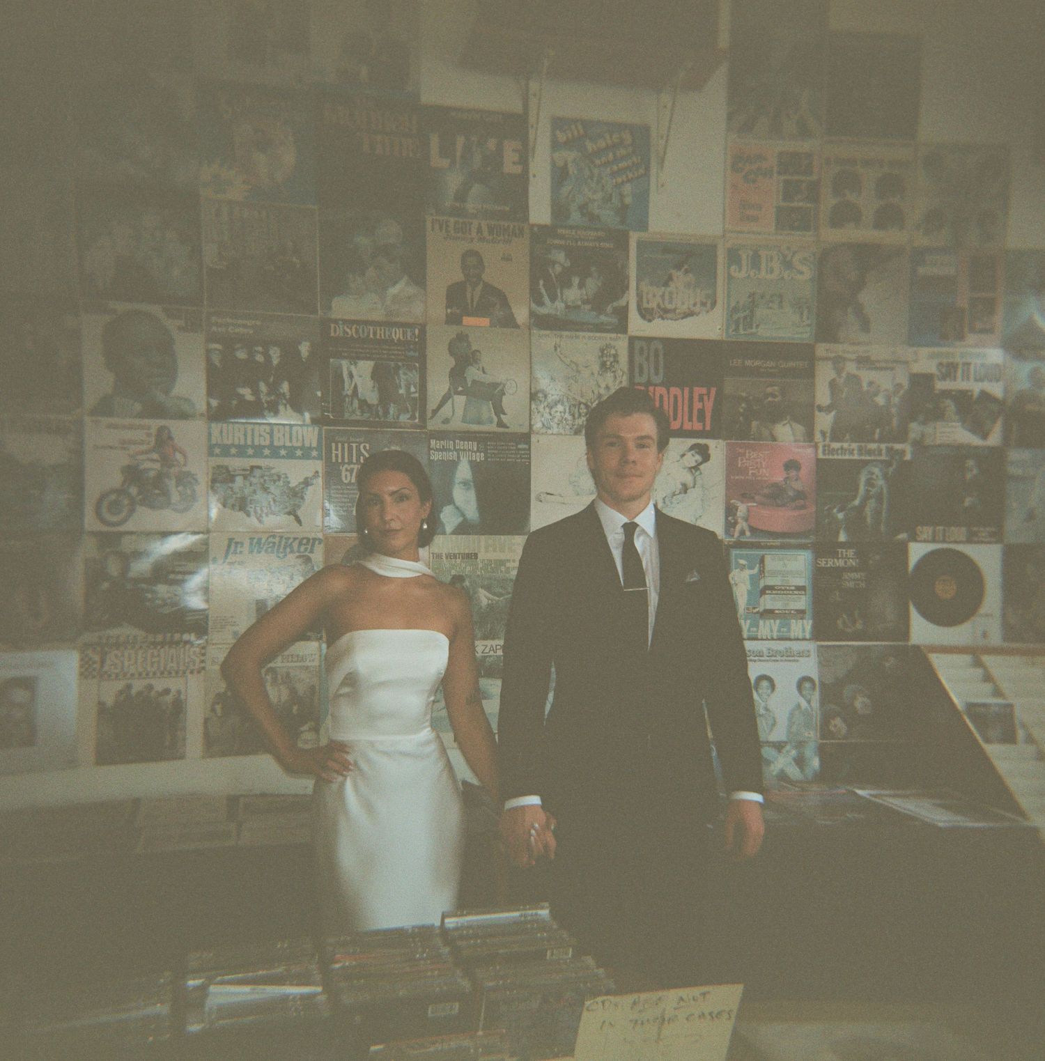 Bride in a white strapless gown and groom in a dark suit stand in a vintage record shop with vinyl album covers lining the walls.