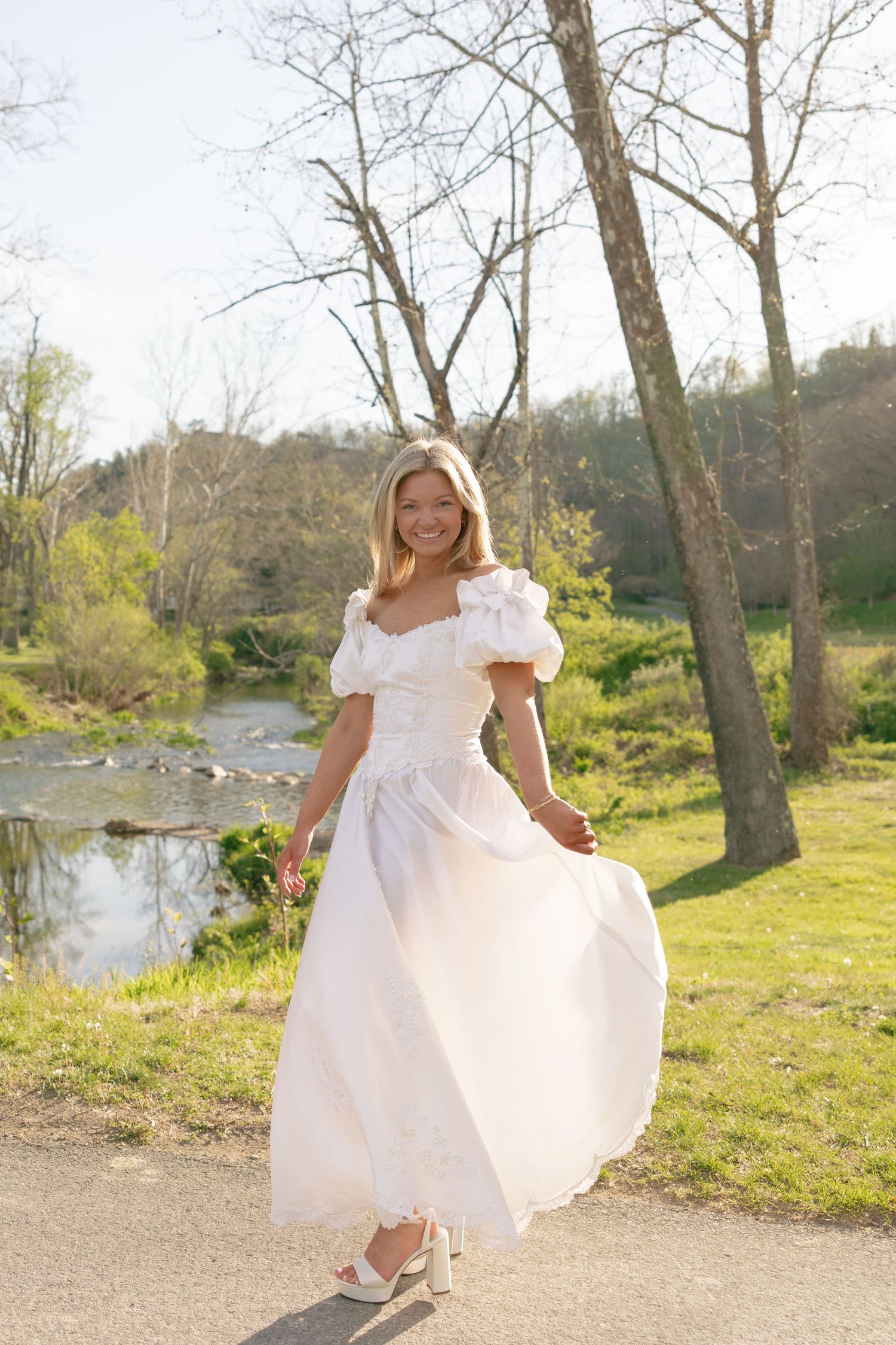 Smiling bride in white puff-sleeve gown twirling outdoors near a serene pond surrounded by trees.