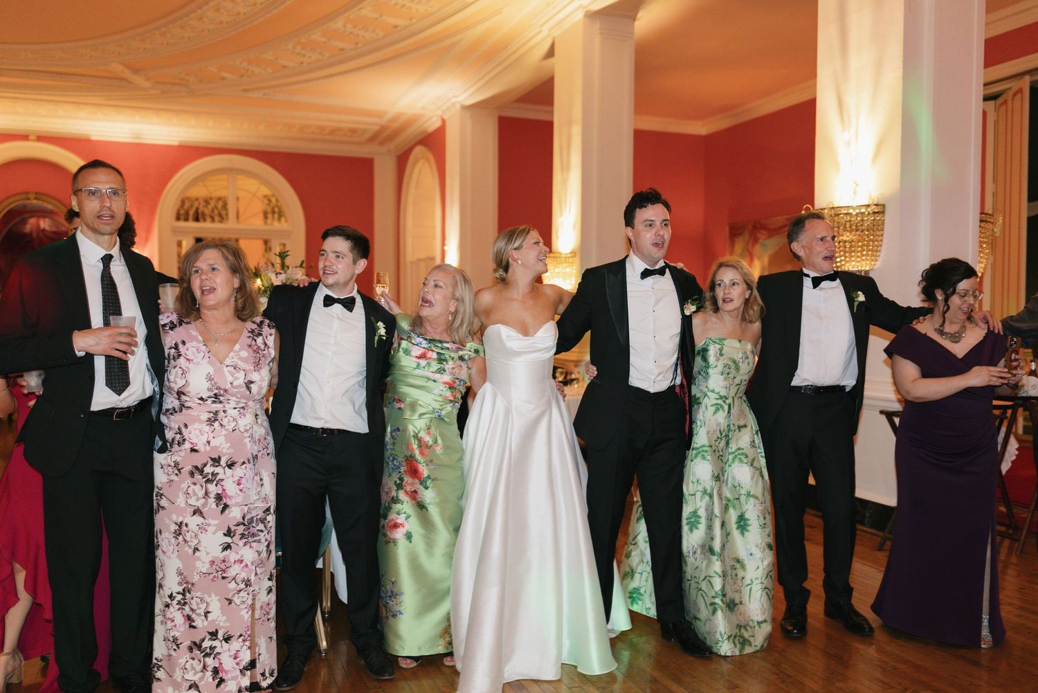 Wedding party in formal attire, bride in white gown, guests in tuxedos and floral gowns, posing in elegant red room.
