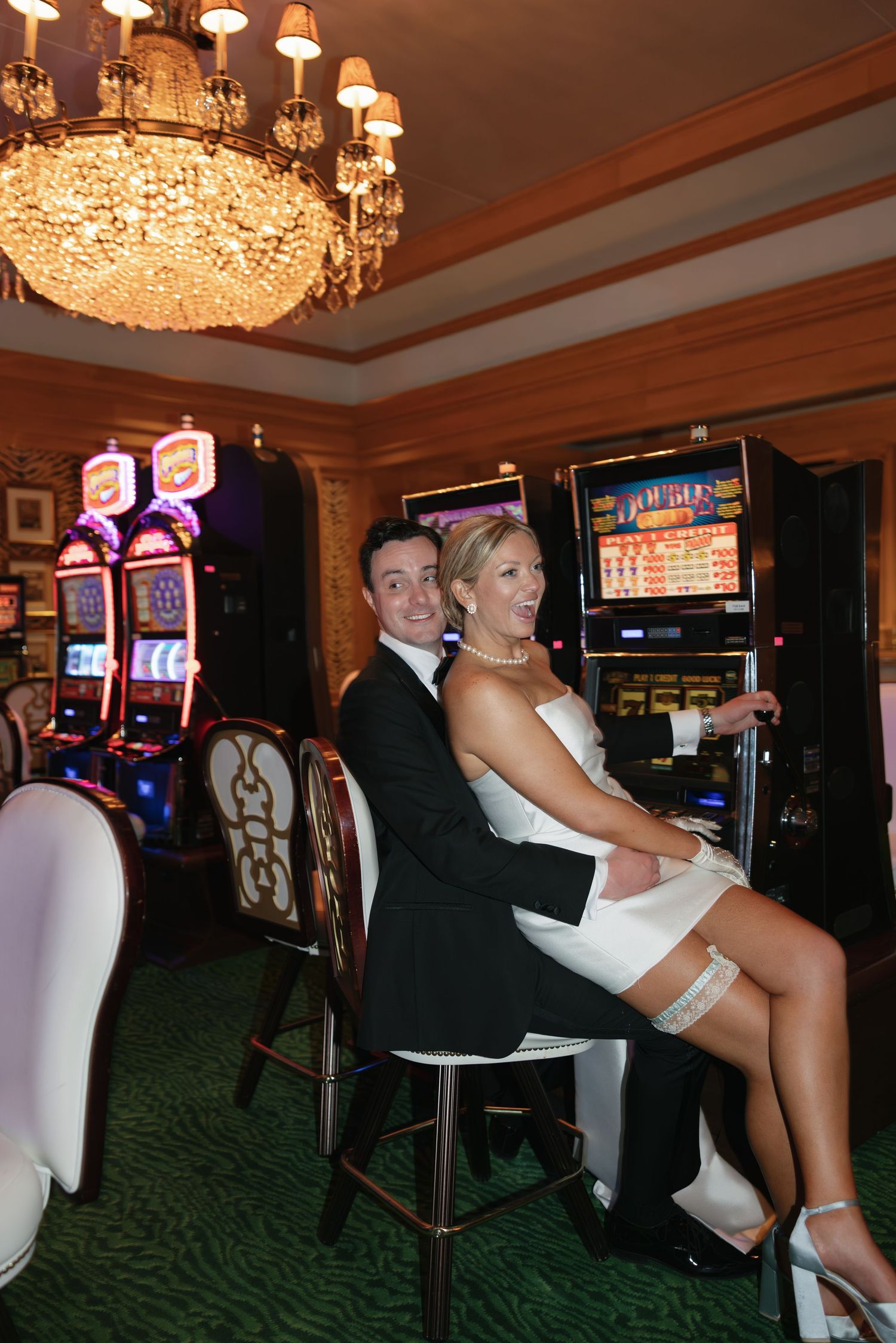 Newlywed couple in wedding attire sitting together at a slot machine in an elegant casino.