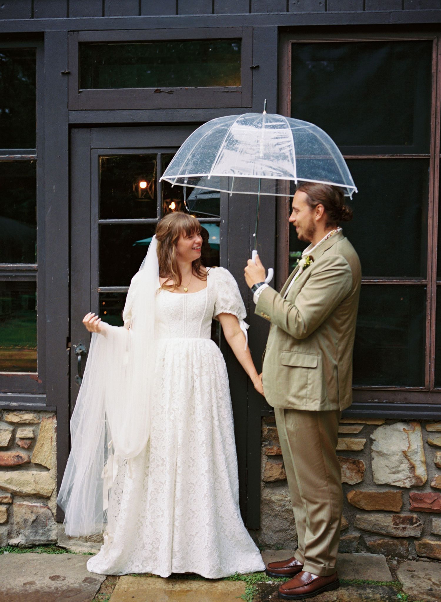 A couple shares a clear umbrella while standing outside a rustic building with stone walls and dark windows.