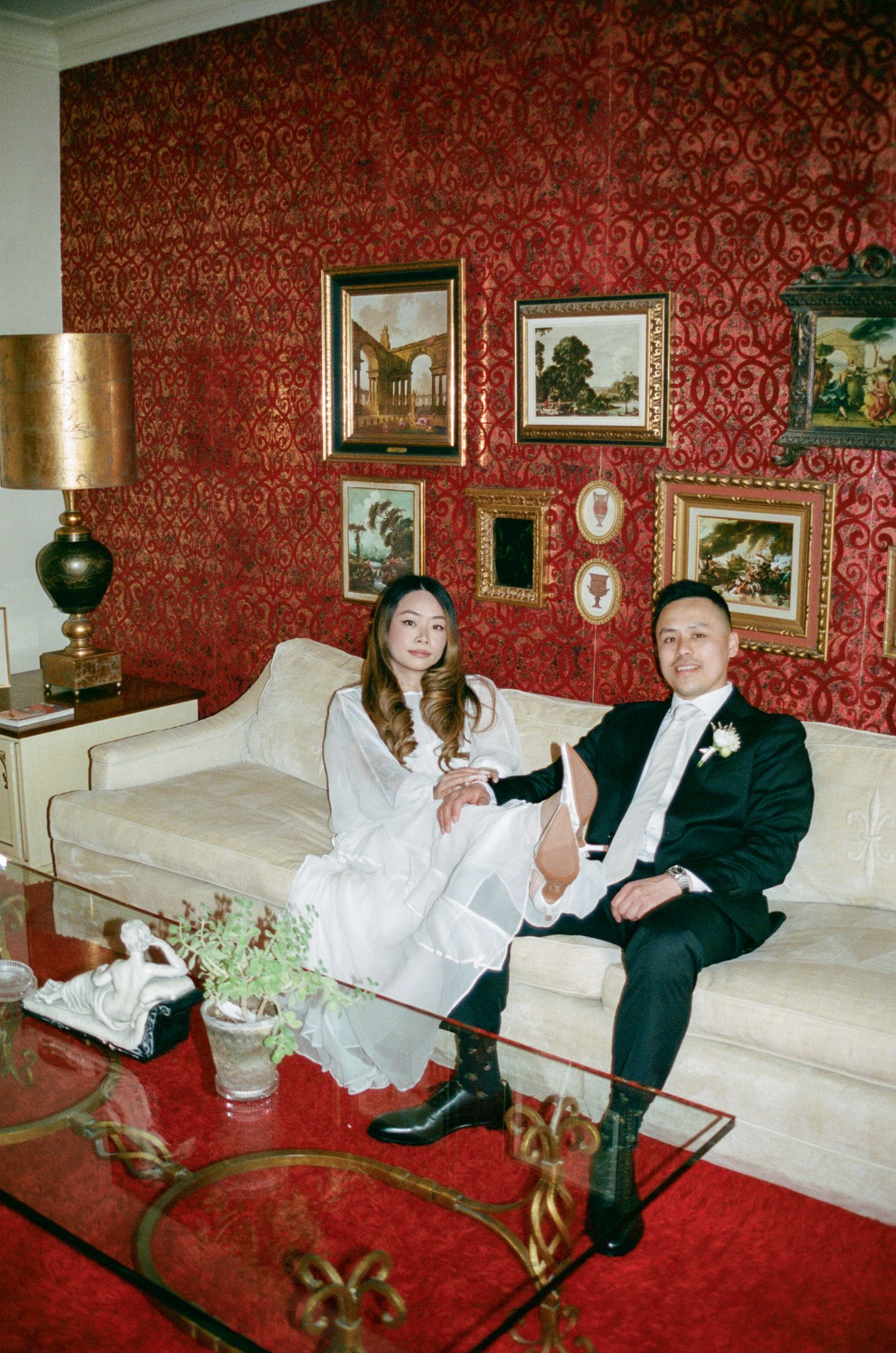 A couple relaxes on a white couch in a formal room with red damask wallpaper and vintage decor.