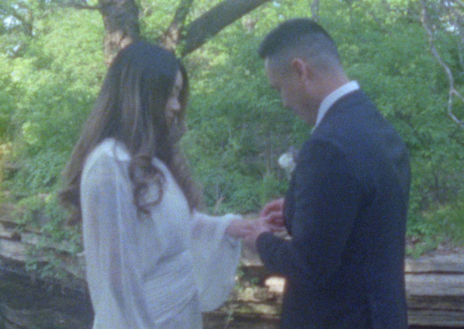 A couple stands together outdoors in a wooded area during what appears to be a romantic moment.