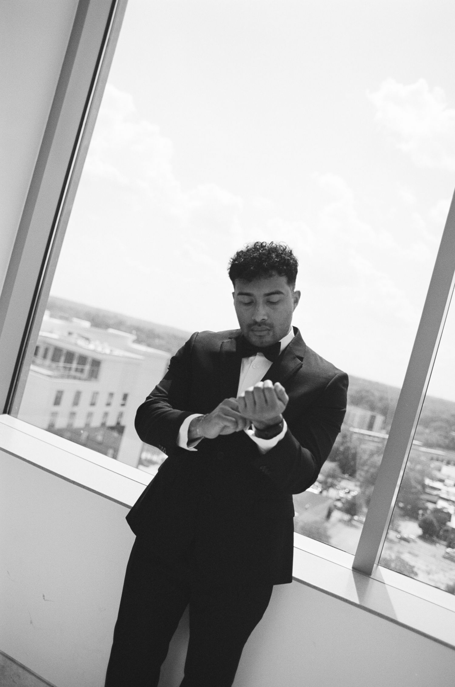 A person in a black suit checks their watch while standing near a large window overlooking a cityscape.
