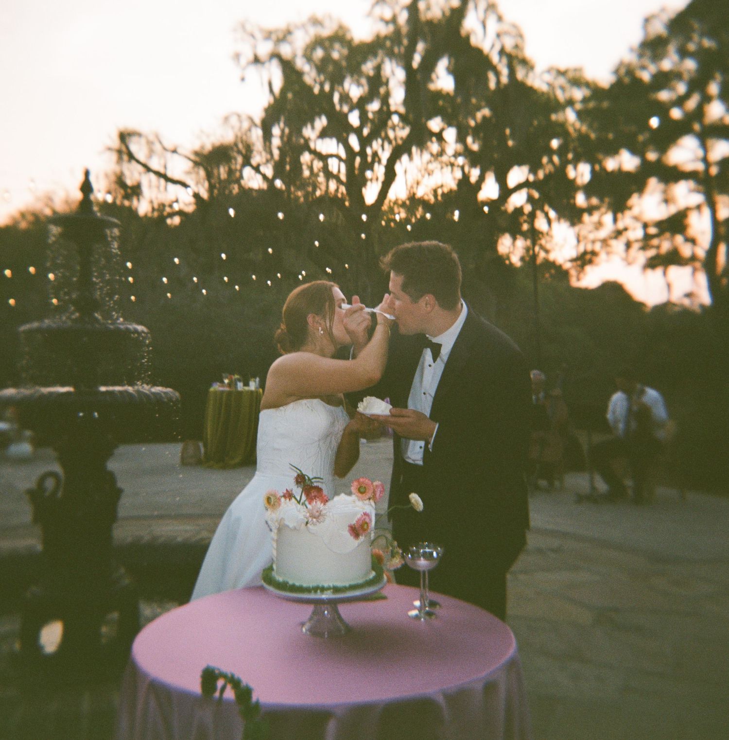 Bride and groom sharing wedding cake at outdoor sunset reception with string lights and trees in background.