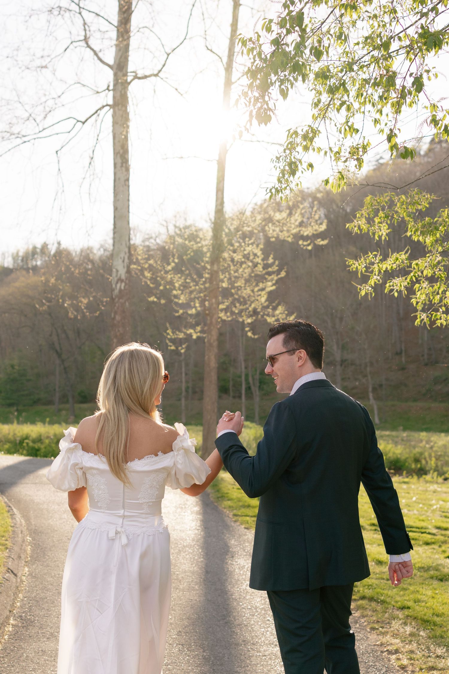 Bride and groom walking hand in hand on a sunlit path surrounded by lush greenery during golden hour.