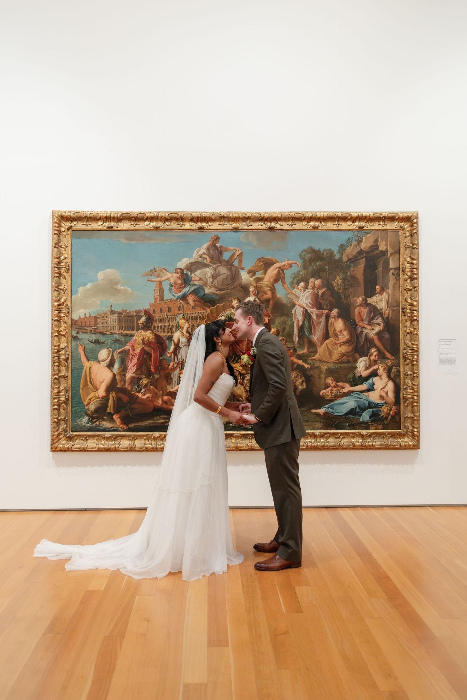 Bride and groom kissing in front of a large ornate painting in an art museum on their wedding day.