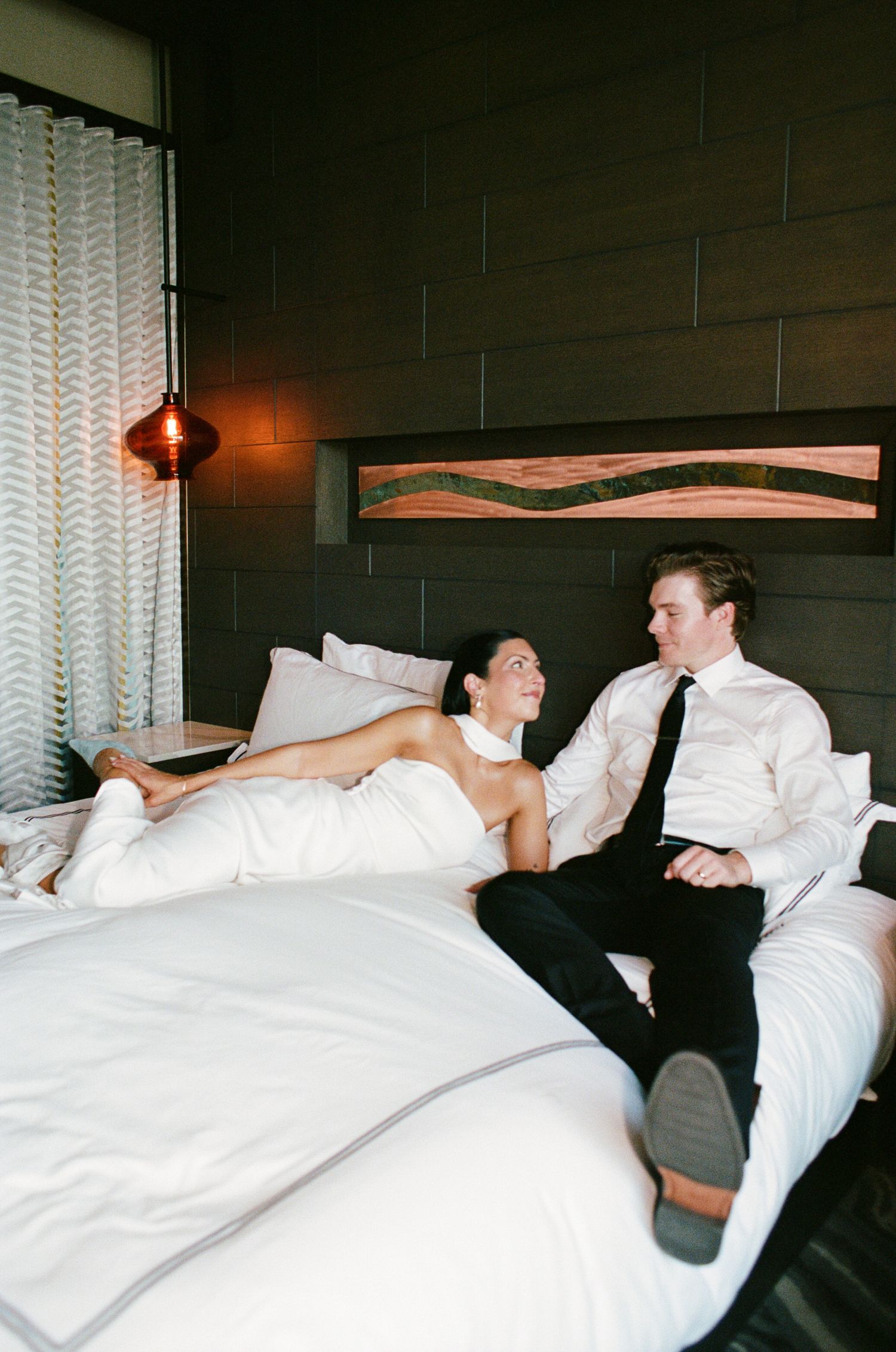 A bride in a white gown and a groom in a white shirt and black tie relax together on a white hotel bed in a modern room.