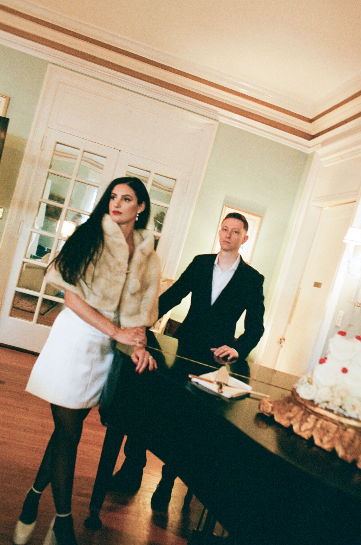 A woman in a white dress and fur stole stands beside a man in a dark suit at a black piano in an elegant room with a decorated cake.