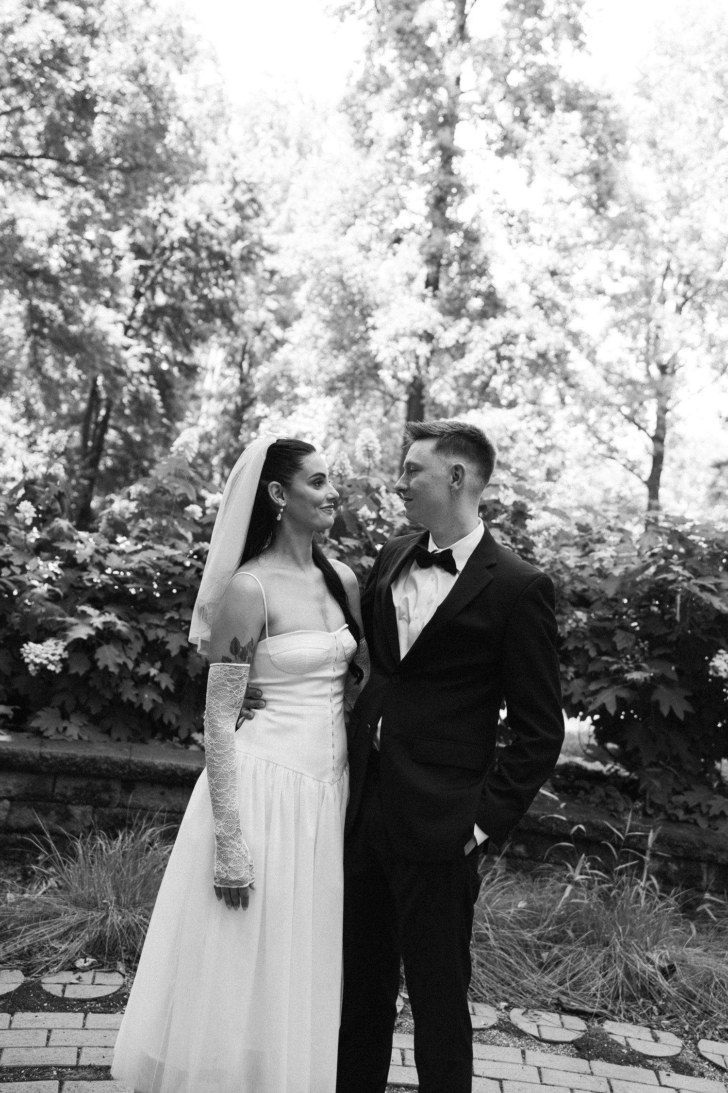Black and white photo of a bride in a veil and long lace glove gazing at a groom in a tuxedo and bow tie in a leafy garden.